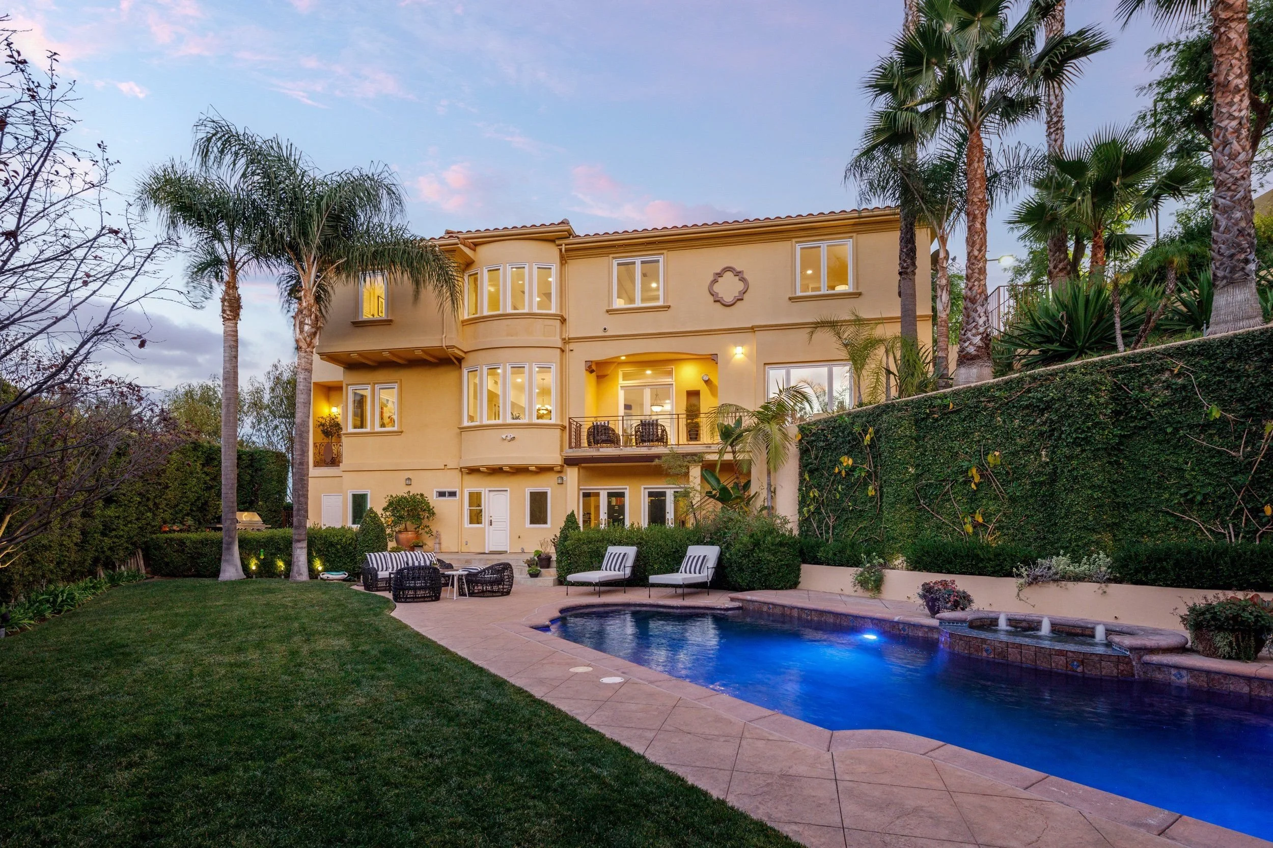 Backyard with a swimming pool, outdoor seating, and a beige multi-story house with several windows and a balcony, surrounded by tall palm trees and lush greenery.