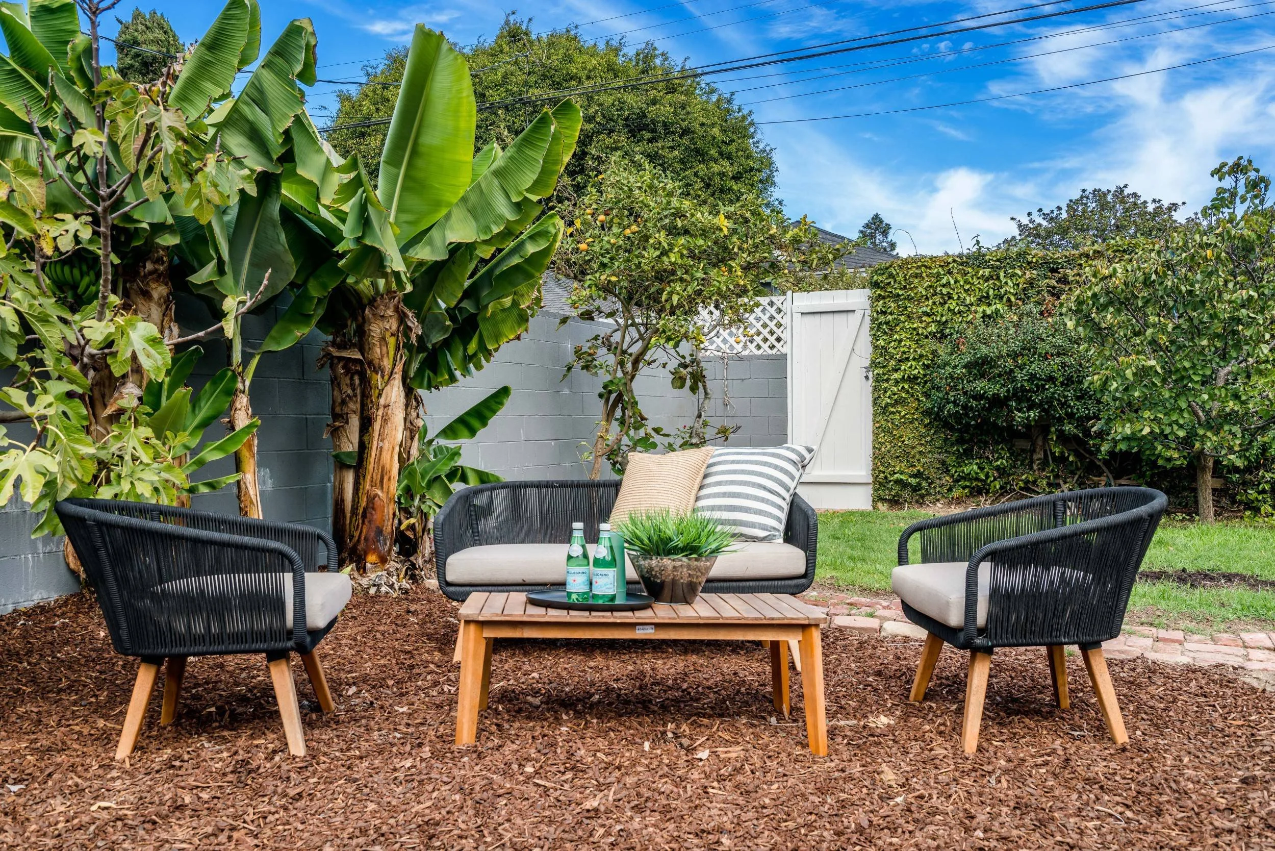 Outdoor patio area with black woven chairs, a wooden coffee table, and a cushioned bench with striped pillows, surrounded by lush greenery including banana plants and trees, with a brick and wood fence under a blue sky with clouds.