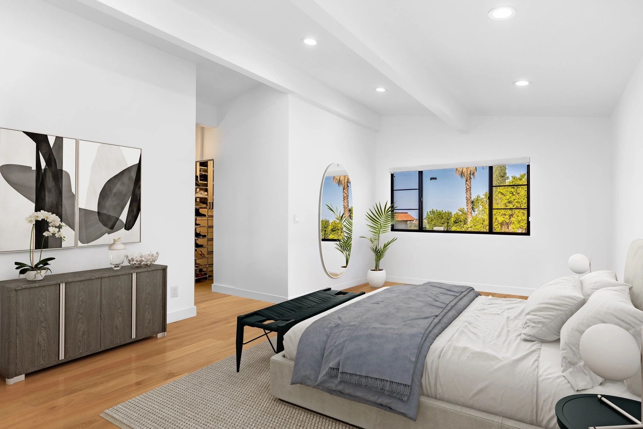 Modern bedroom with white walls, large window showing trees outside, a bed with gray and white bedding, a wooden dresser with decorative items, and minimalist artwork on the wall.