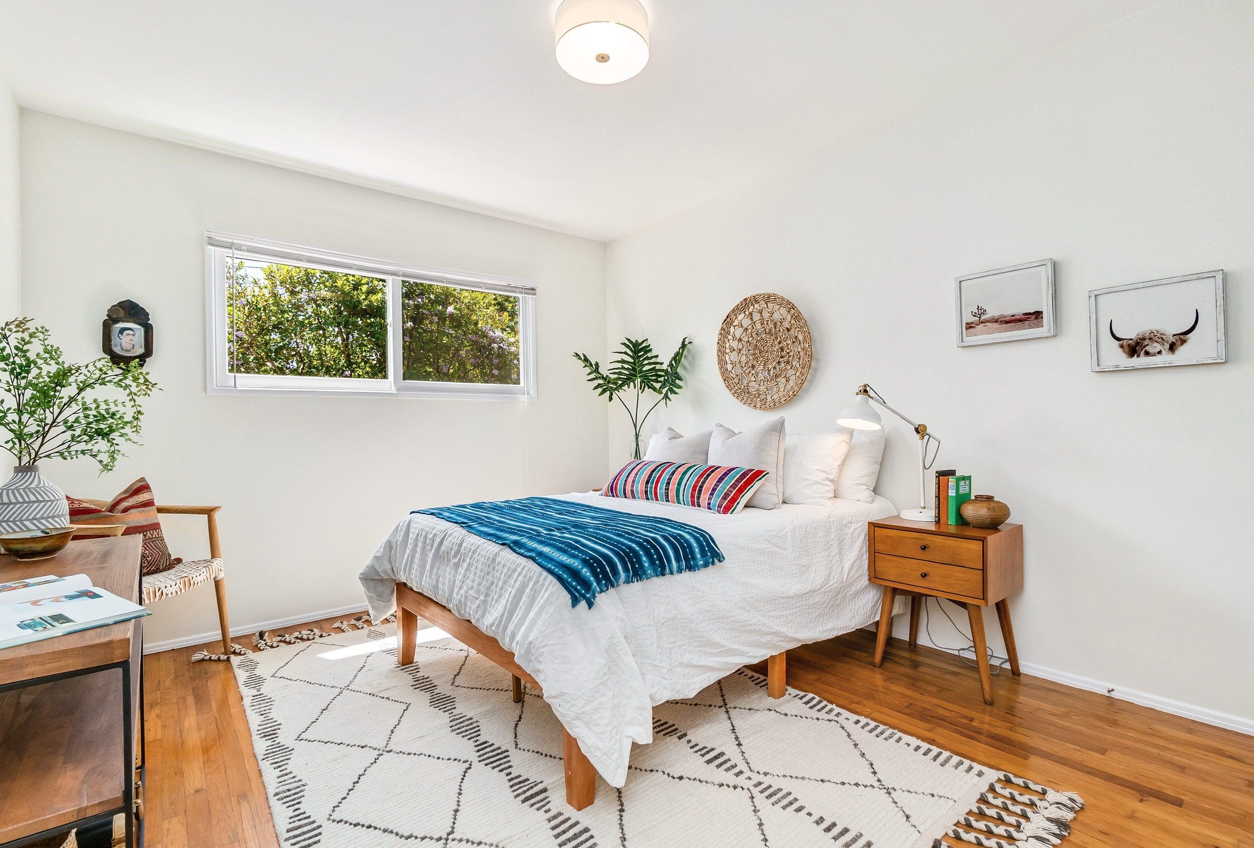 A bedroom with white walls, a wooden bed with white bedding and colorful pillows, a bedside table with a lamp and books, wall art including a photo of a cow with horns and landscape, a woven wall decor, a dresser, plants, and a window with greenery o