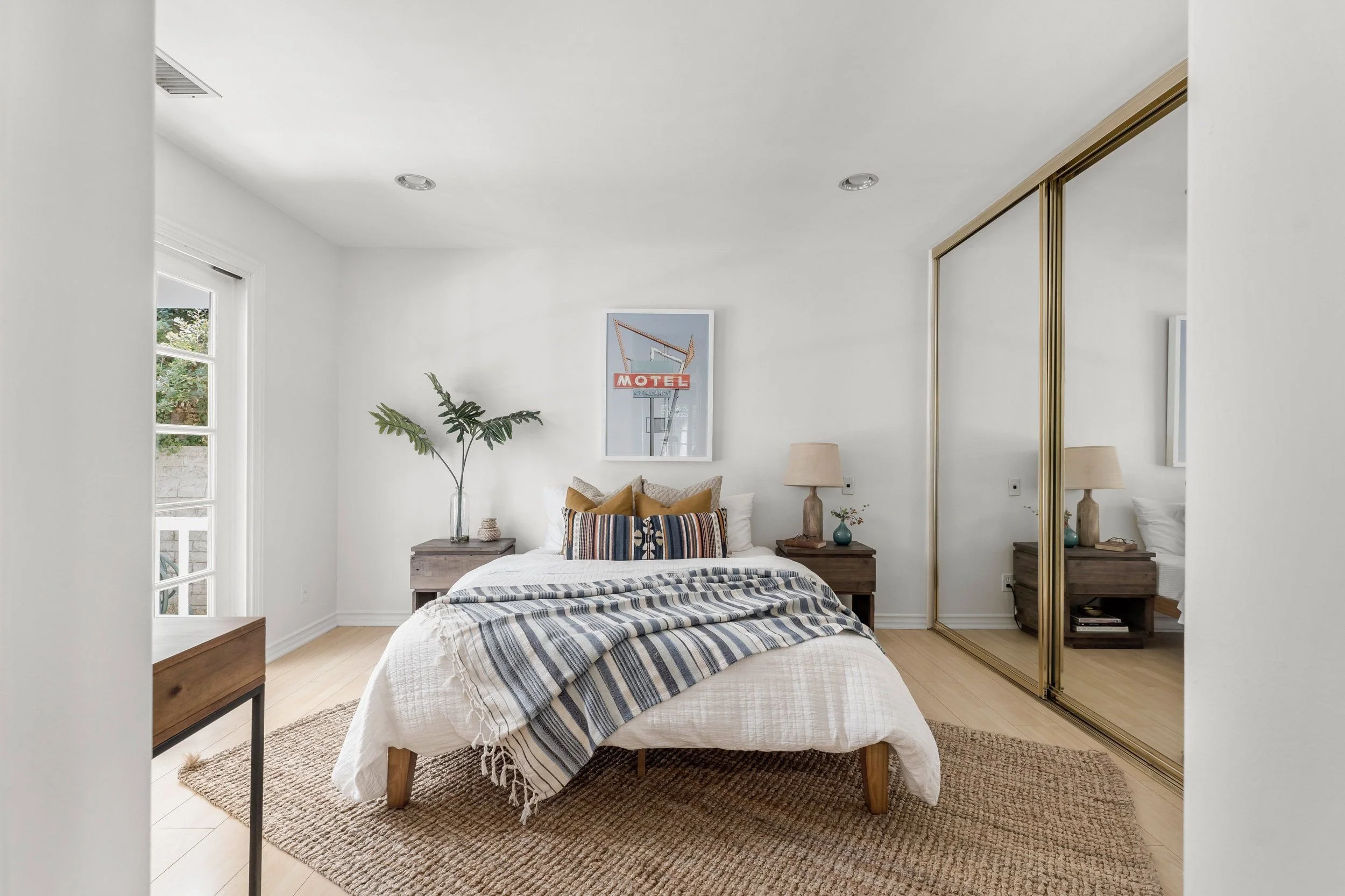 Bedroom with white walls and ceiling, a bed with striped bedding and pillows, two bedside tables with lamps, a framed motel sign poster on the wall, a plant in a clear vase, a window with greenery outside, and mirrored closet doors reflecting part of