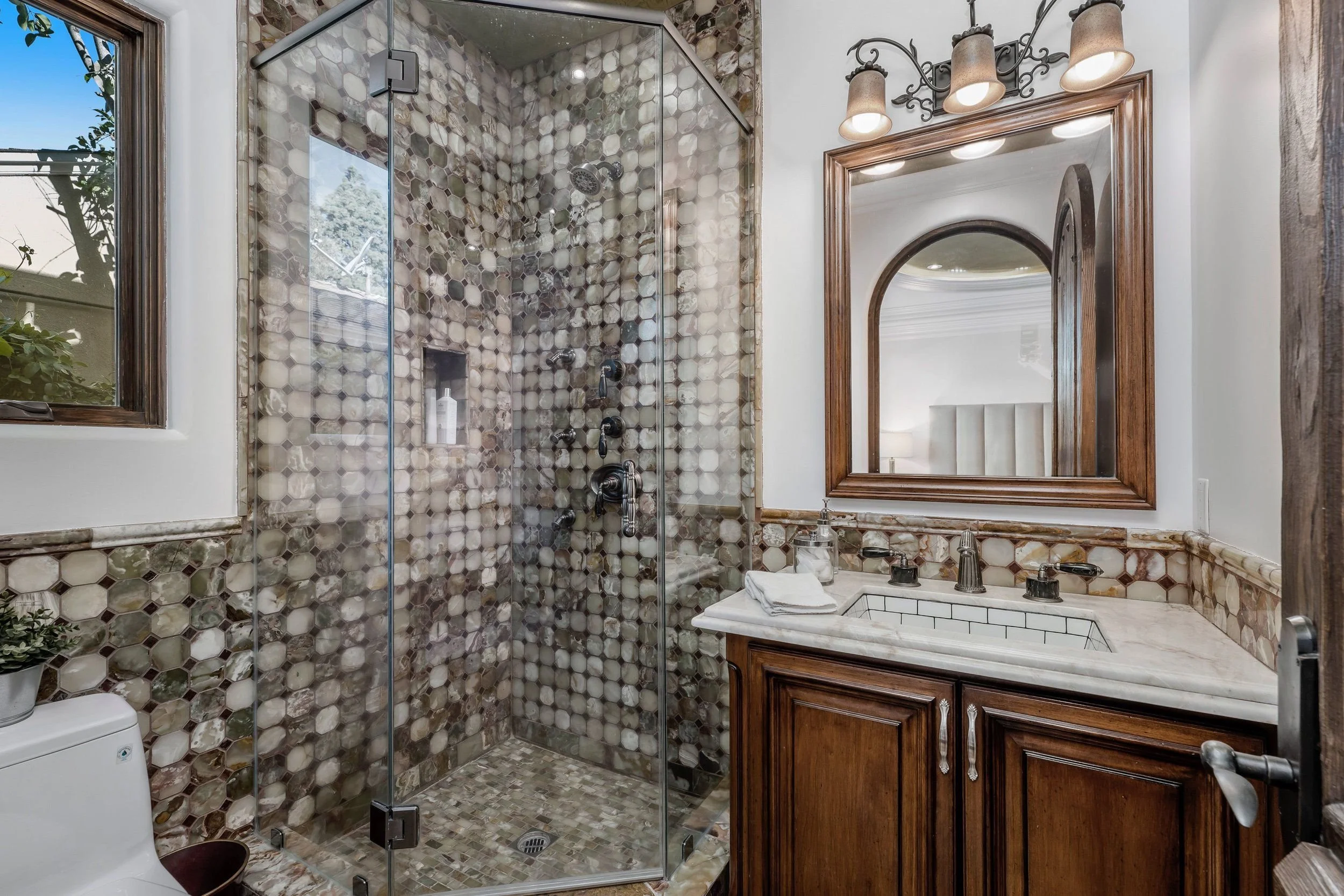 Bathroom with a glass-enclosed shower featuring hexagonal and marble tile pattern, a mirror framed in wood above a marble countertop with a sink, and a window showing outdoors. There are light fixtures above the mirror and a toilet partially visible 