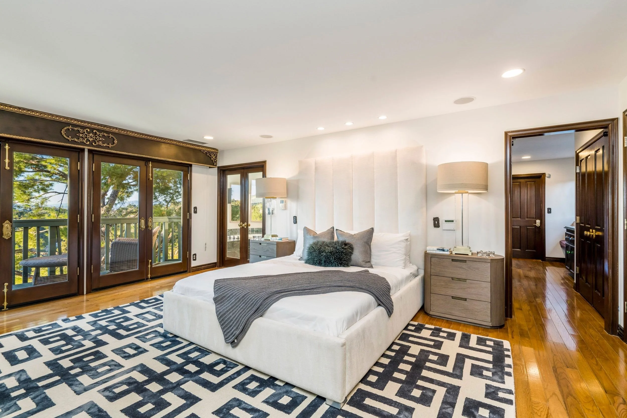 A bedroom with a large bed decorated with pillows, a throw blanket, and a geometric rug. The room has wooden windows, a nightstand, and tall white lamps. There are multiple ceiling lights and a door leading to another room.