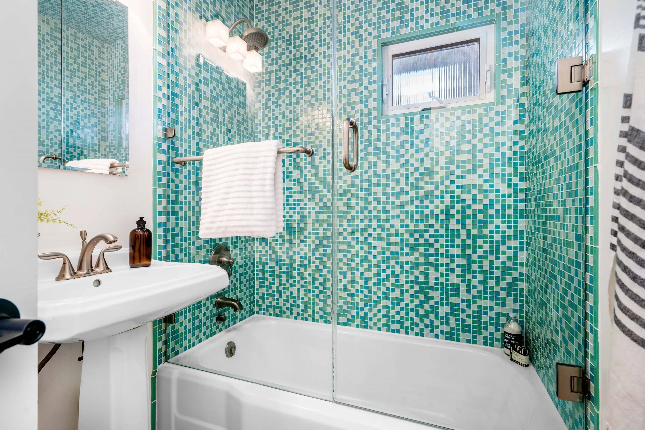A bathroom with green and blue mosaic tiles on the walls, a glass shower door, a white bathtub, a white towel over a grab bar, a small window, a white sink with bronze fixtures, a soap dispenser, and bottles of bath products.
