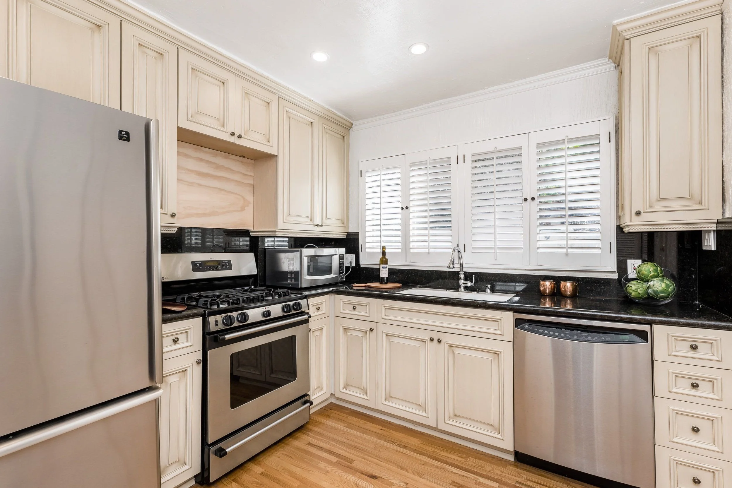 A kitchen with beige cabinets, black granite countertops, stainless steel appliances including a refrigerator and stove, and three small copper cups on the counter. There are three bowls of green artichokes on the right side of the counter, and a bot