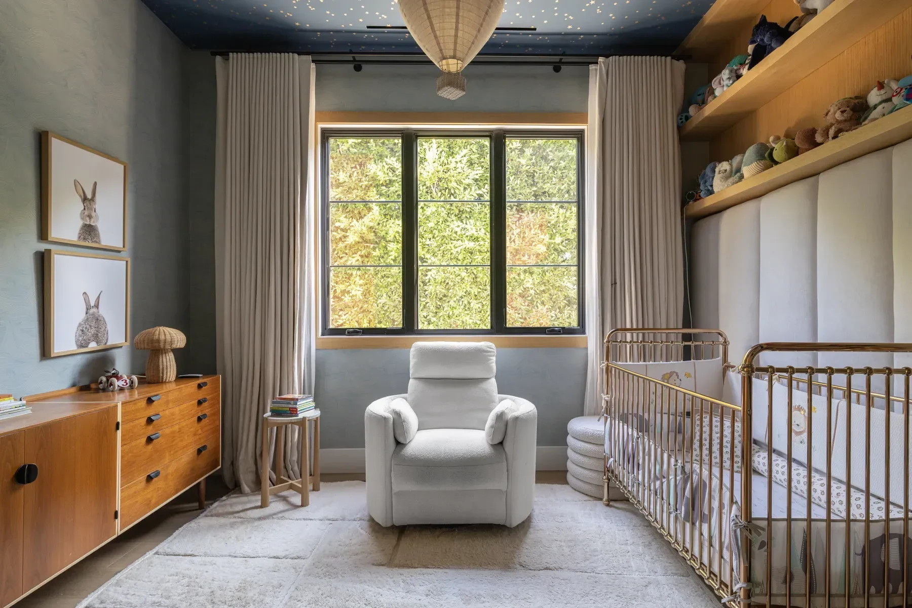 A cozy diaper-changing room with a large window showing trees outside. The room has a white armchair in the center, a wooden dresser with toys and books, and a gold metal crib with baby bedding. Decorative art of rabbits and a starry ceiling add a wh