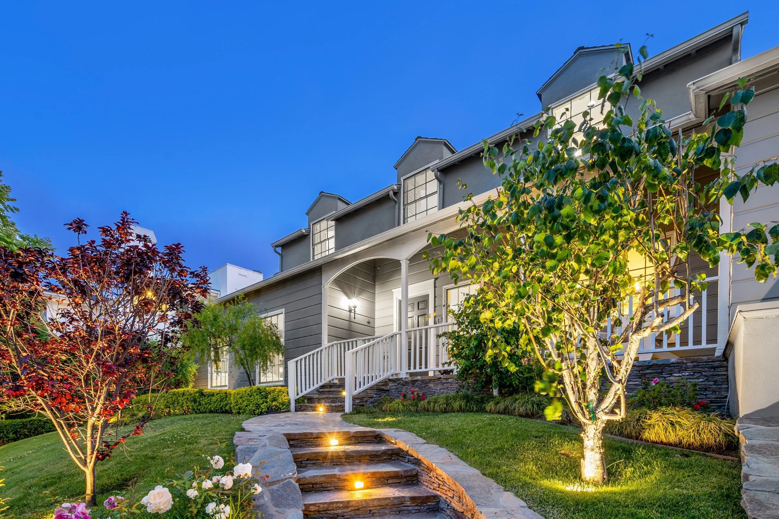 A well-lit front yard garden with steps leading to a house, a large tree, colorful flowers, shrubs, and greenery under a dusk sky.
