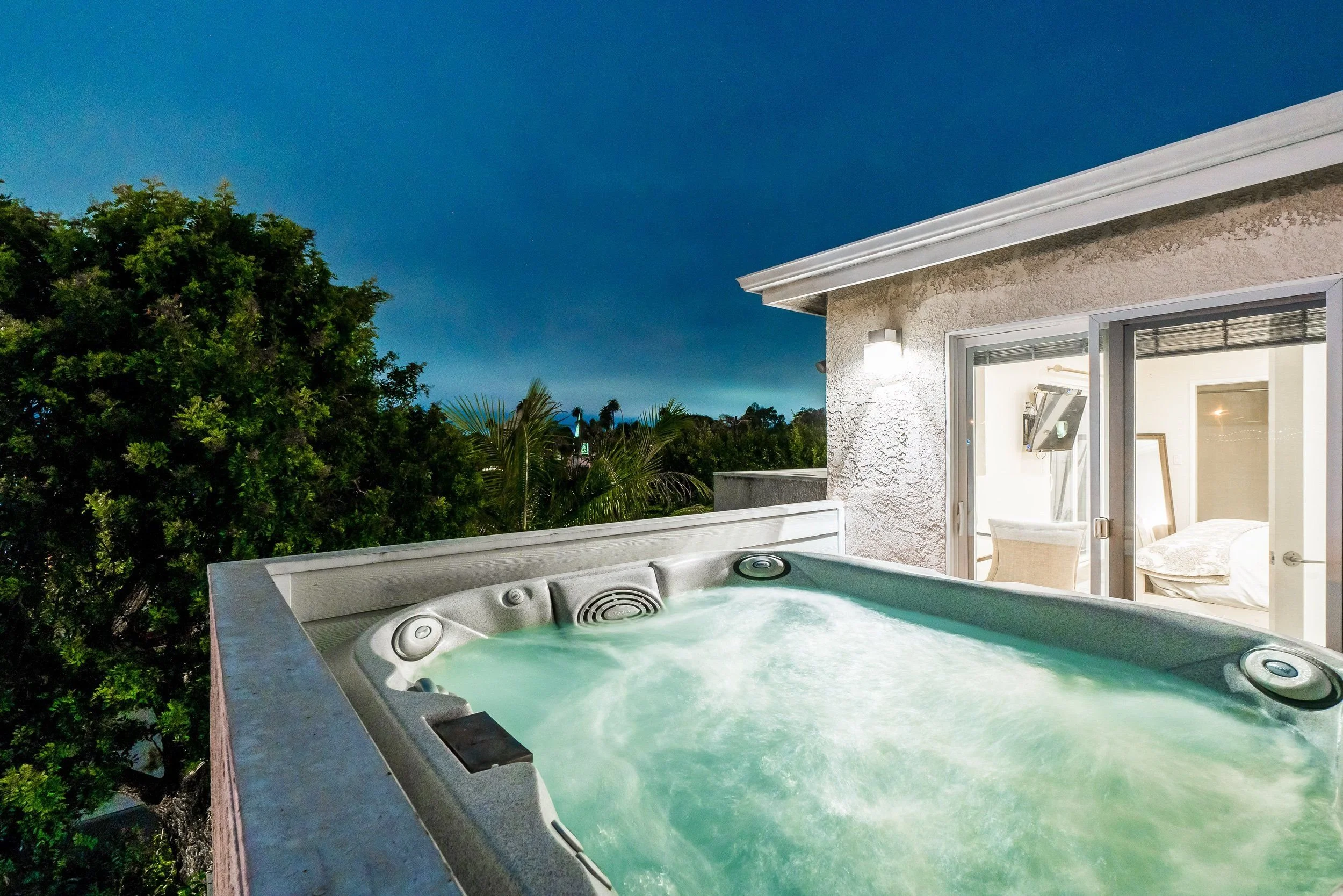 An outdoor hot tub on a balcony with a view of trees and a blue sky, part of a residential home with an open sliding door revealing a bedroom inside.