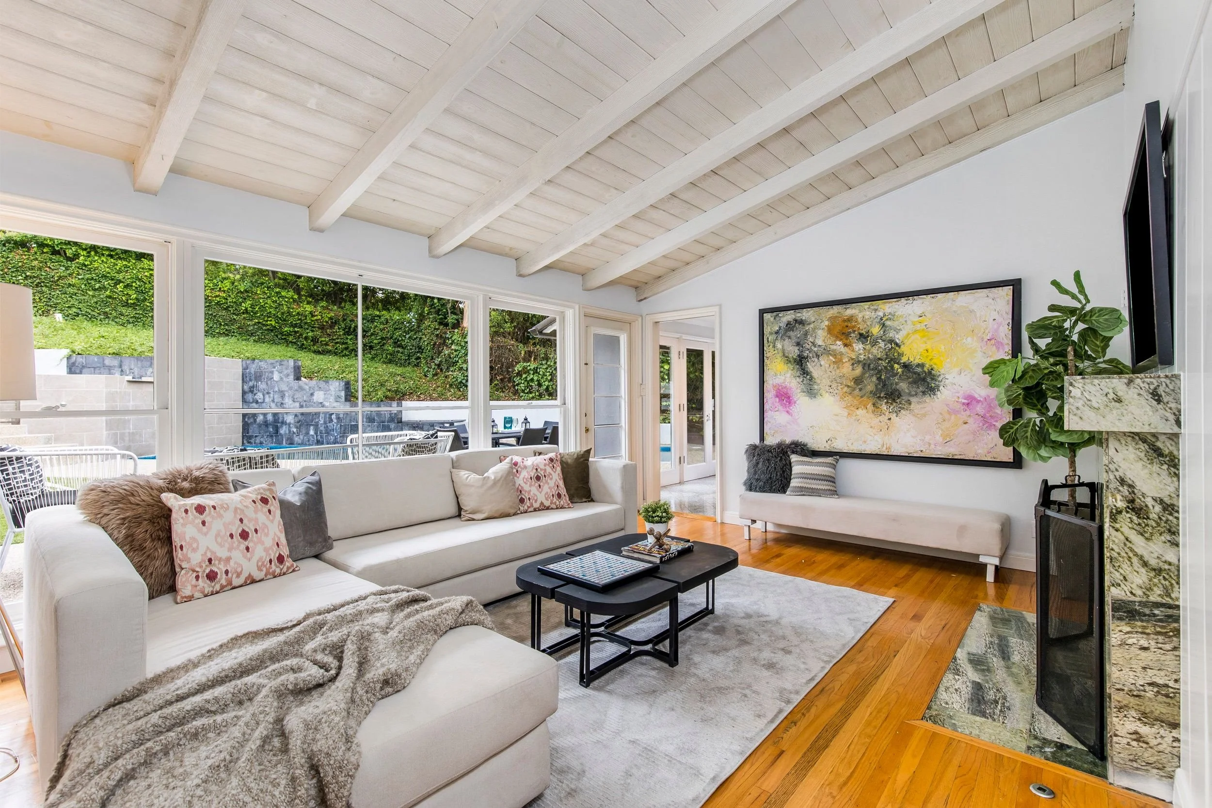 Living room with white sofa, coffee table, large window showing outdoor patio and greenery, wall art, potted plant, and a corner fireplace.