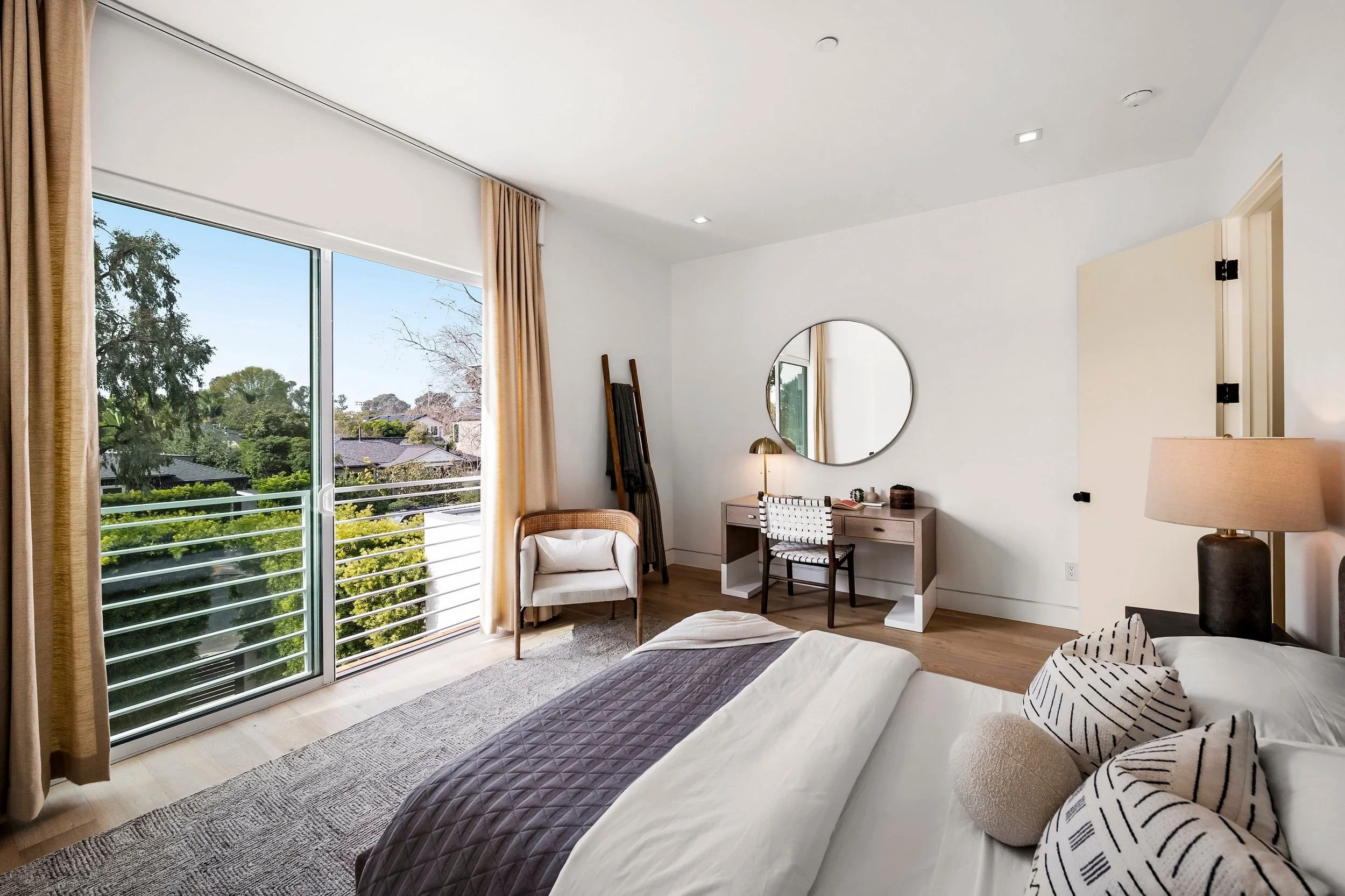 A modern bedroom with a view outside through a large sliding glass door. The room has beige curtains, a white desk with a round mirror above, a chair, a small table with a gold lamp, and a bed with black and white striped pillows.