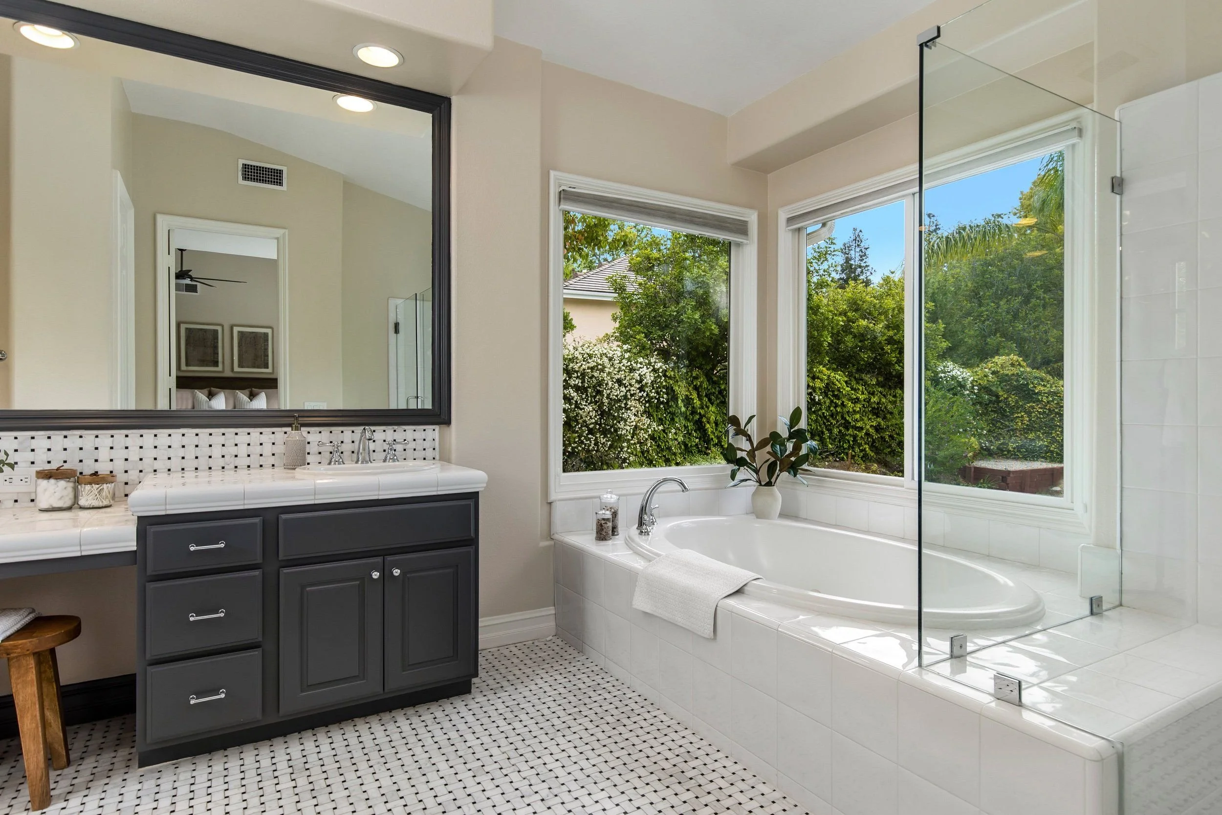 Bathroom with a large window showing greenery outside, a white bathtub, a dark-colored vanity with a white tiled countertop, and a mirror reflecting a bedroom door and artwork.