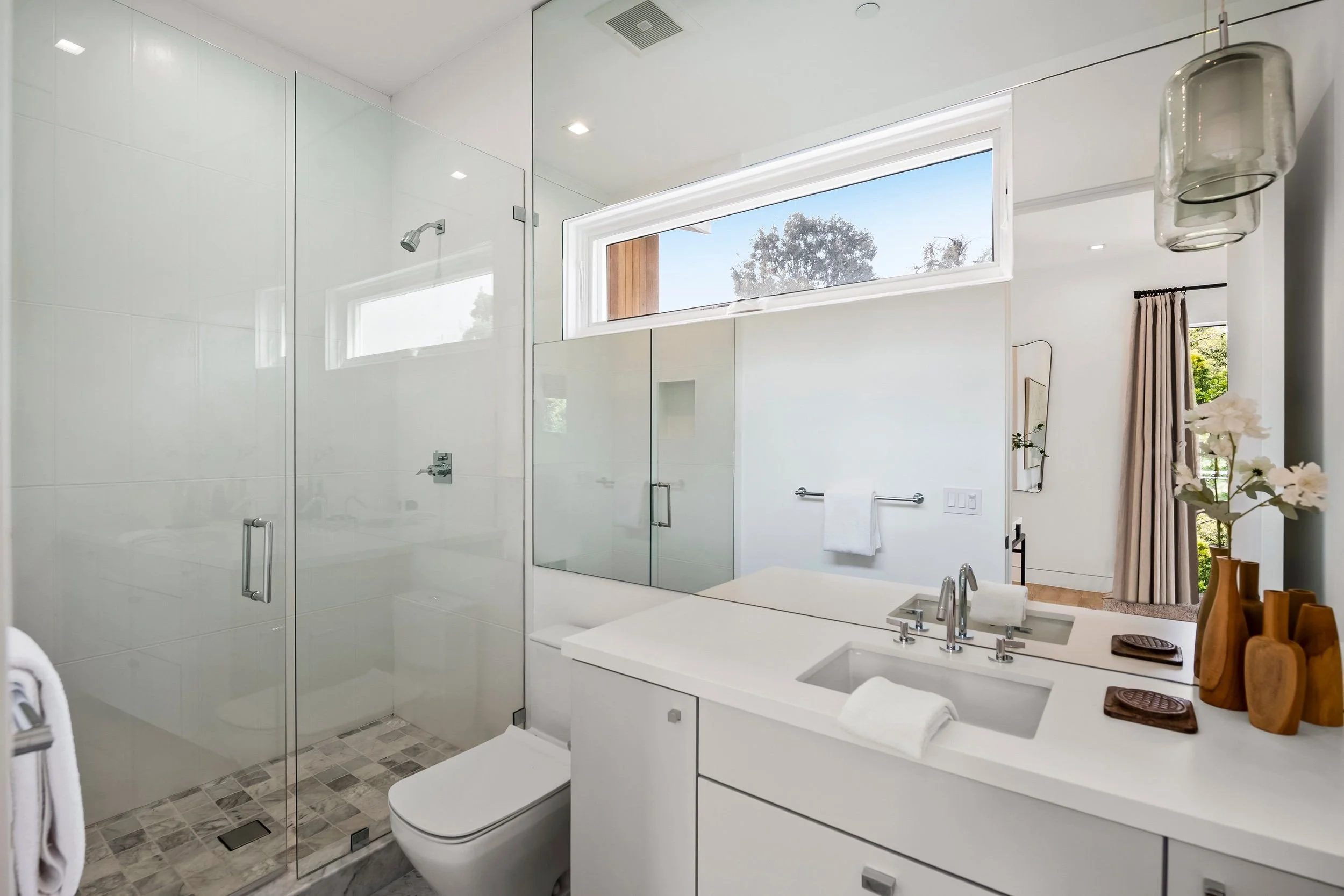 Modern bathroom with a glass shower, white vanity, and large horizontal window showing trees outside. Decor includes a vase with white flowers and wooden accessories.