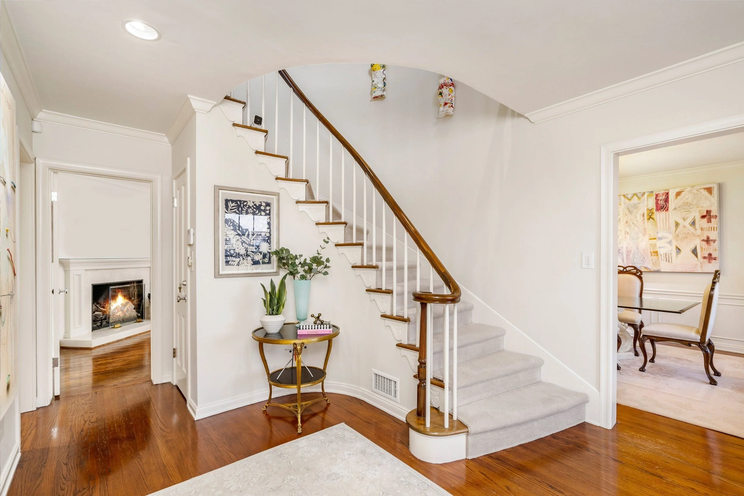 Interior view of a home with a staircase, a fireplace, and a hallway, featuring hardwood floors, white walls, and decorative plants.