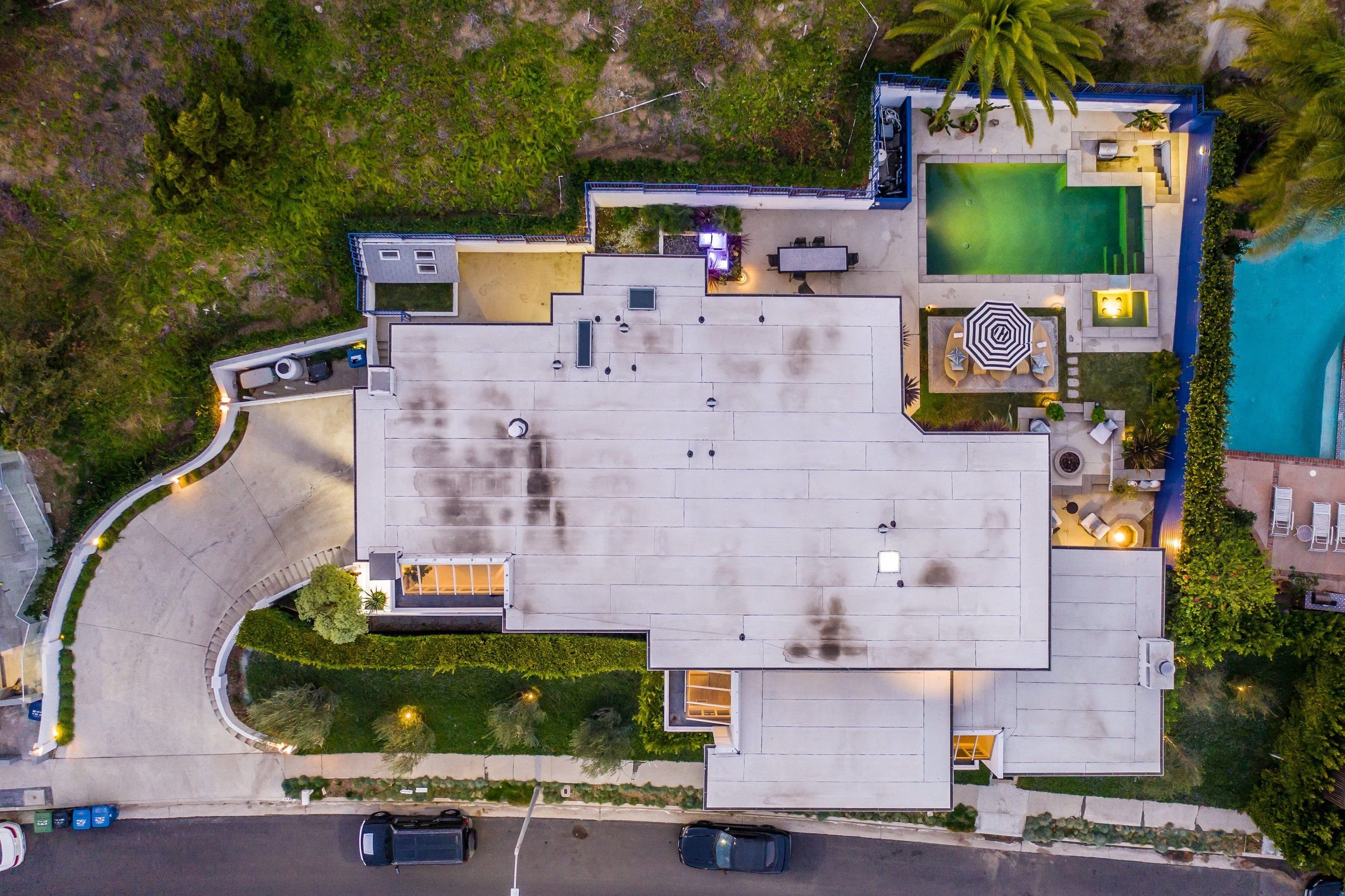 An aerial view of a modern house with a backyard featuring a small pool, outdoor dining area with umbrella, palm trees, and surrounding greenery at dusk.