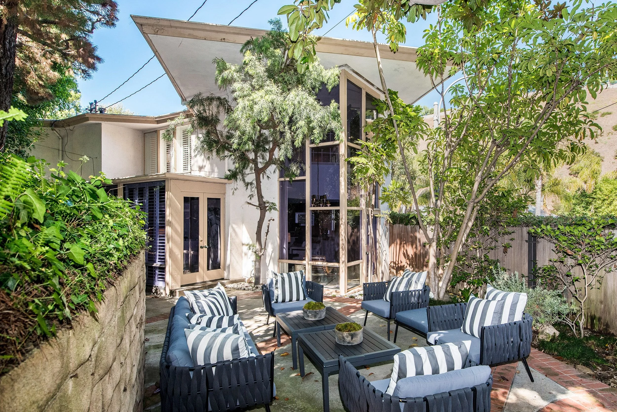 A backyard patio with outdoor seating consisting of cushioned chairs with black and white striped pillows arranged around a rectangular black coffee table. The space is surrounded by trees and lush greenery, with a modern house featuring large window