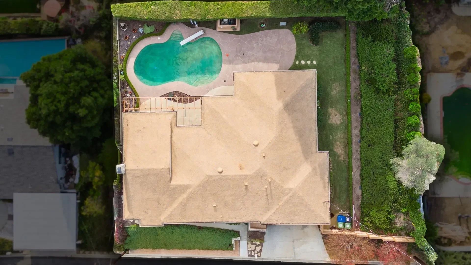 Aerial view of a backyard with a beige house, a stone patio, a small kidney-shaped swimming pool, and surrounding greenery including trees and bushes.