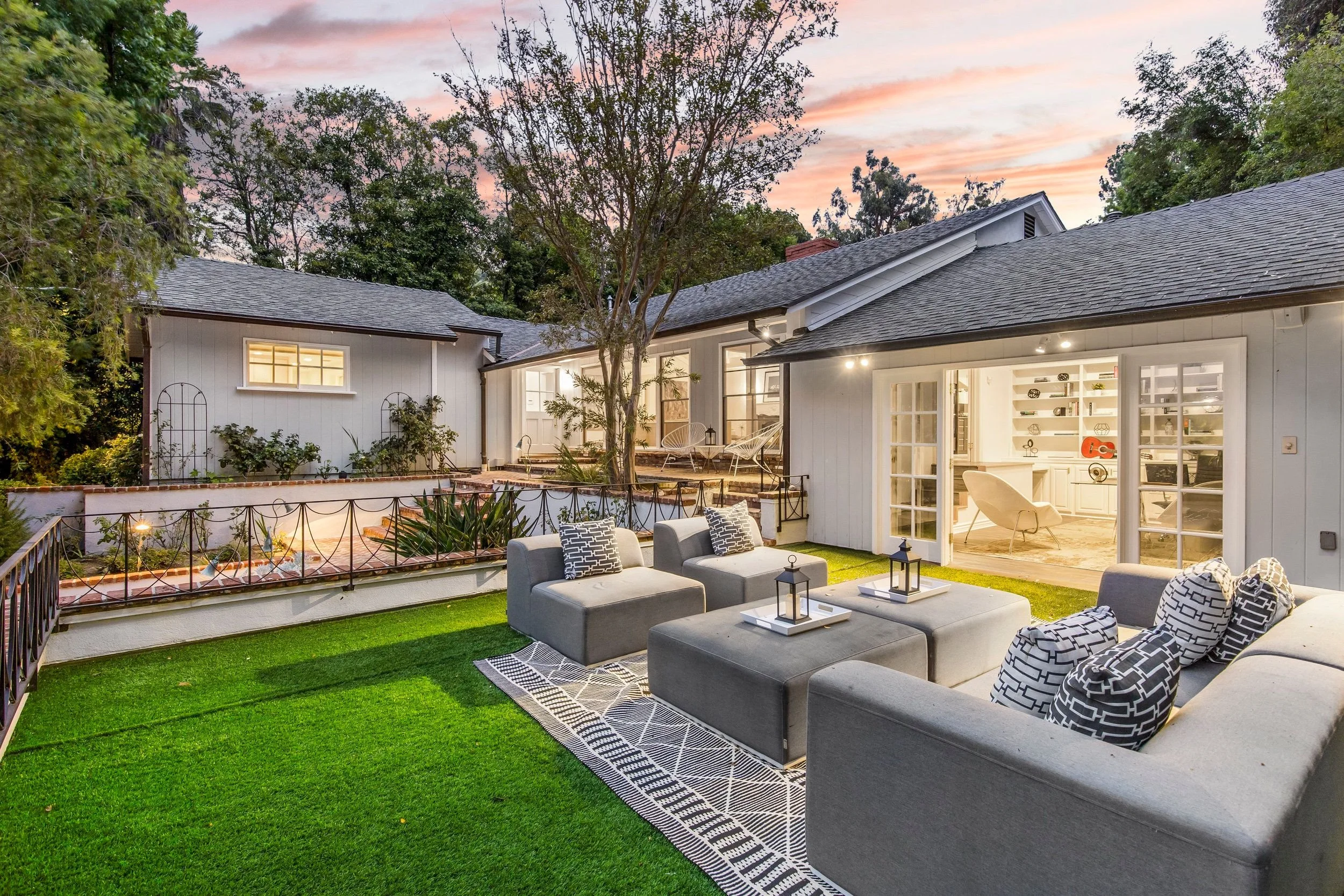 Backyard patio with outdoor seating, a lawn, and a view of a house interior through open sliding doors, during sunset with a colorful sky.