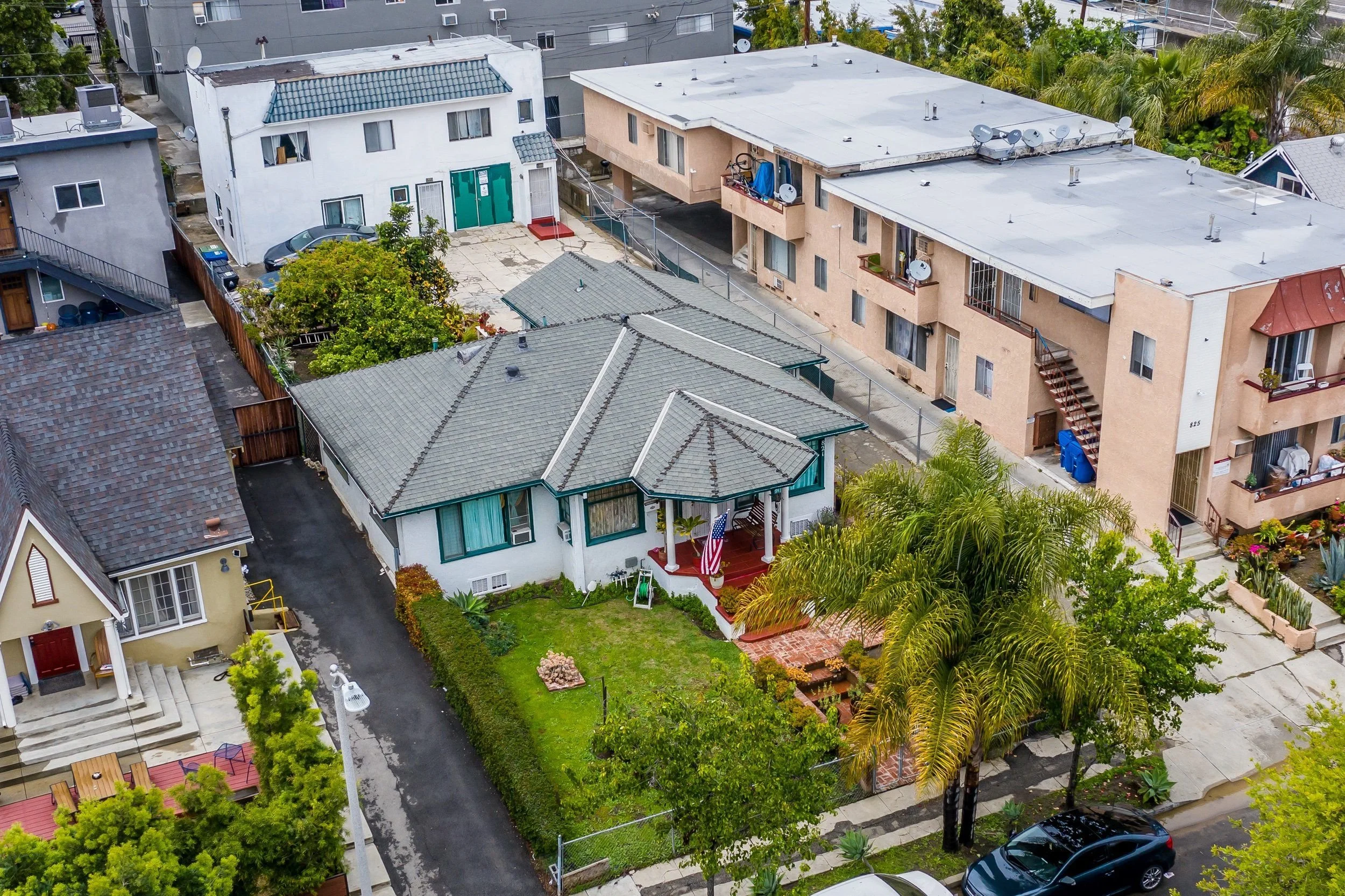 An aerial view of urban residential buildings, including houses with gray tiled roofs, an apartment complex with satellite dishes on its flat roof, a small fenced yard with trees, and various parked cars and greenery.