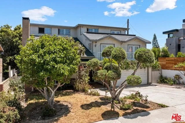 A multi-story residential house with a front yard featuring shaped trees and shrubs, a driveway, and a wooden fence on the right, under a clear blue sky.
