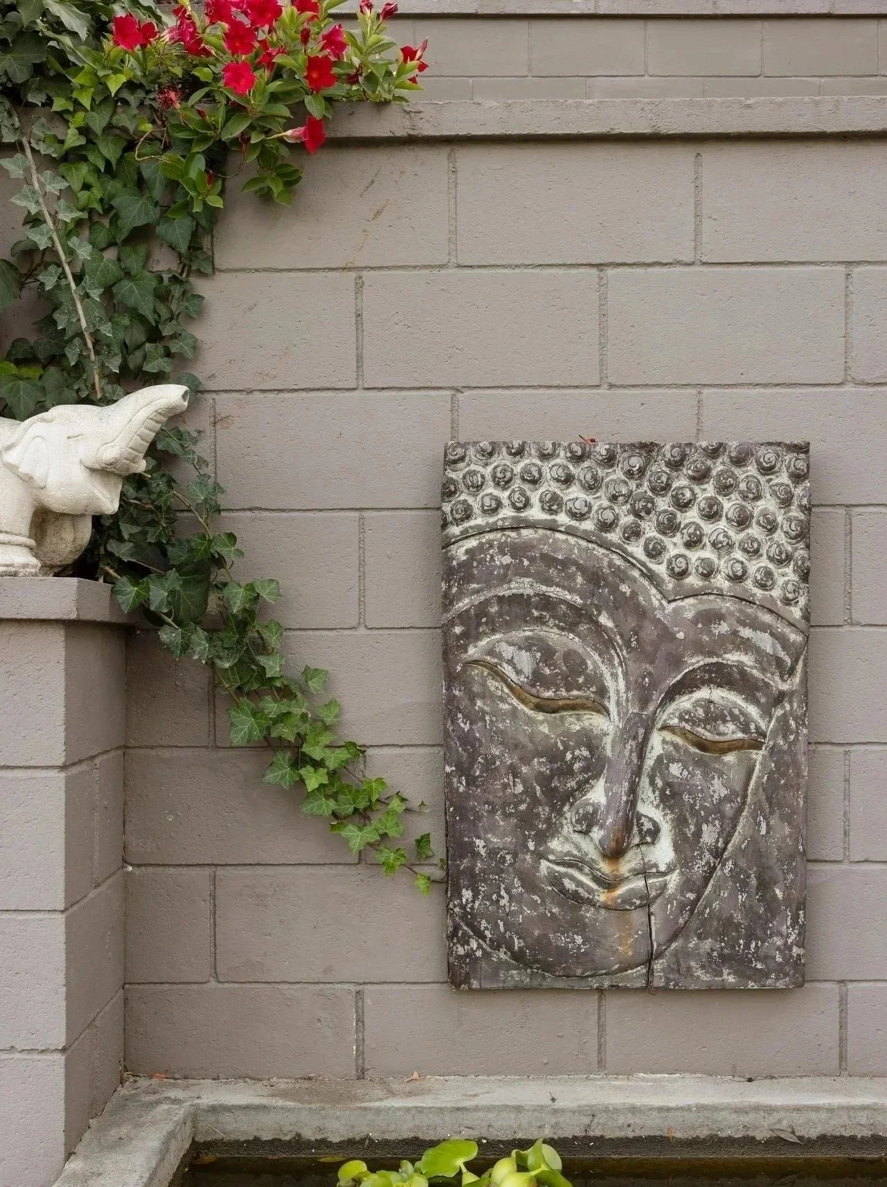 Decorative garden wall with a Buddha face sculpture, green ivy plant, a white ceramic elephant figurine, and water with floating lily pads.
