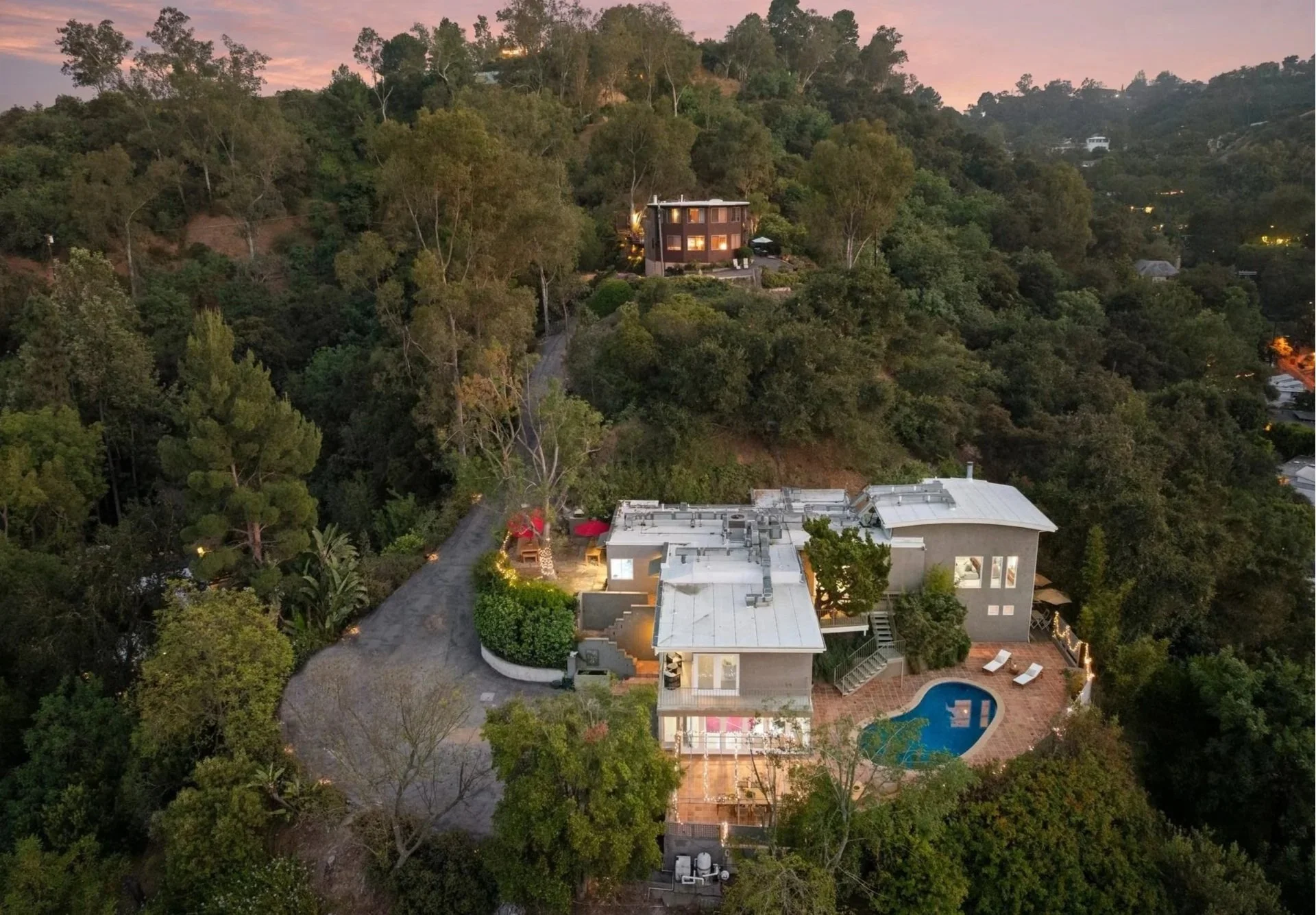 An aerial view of a hillside residential area at dusk showing illuminated houses surrounded by trees, a swimming pool, and outdoor seating.