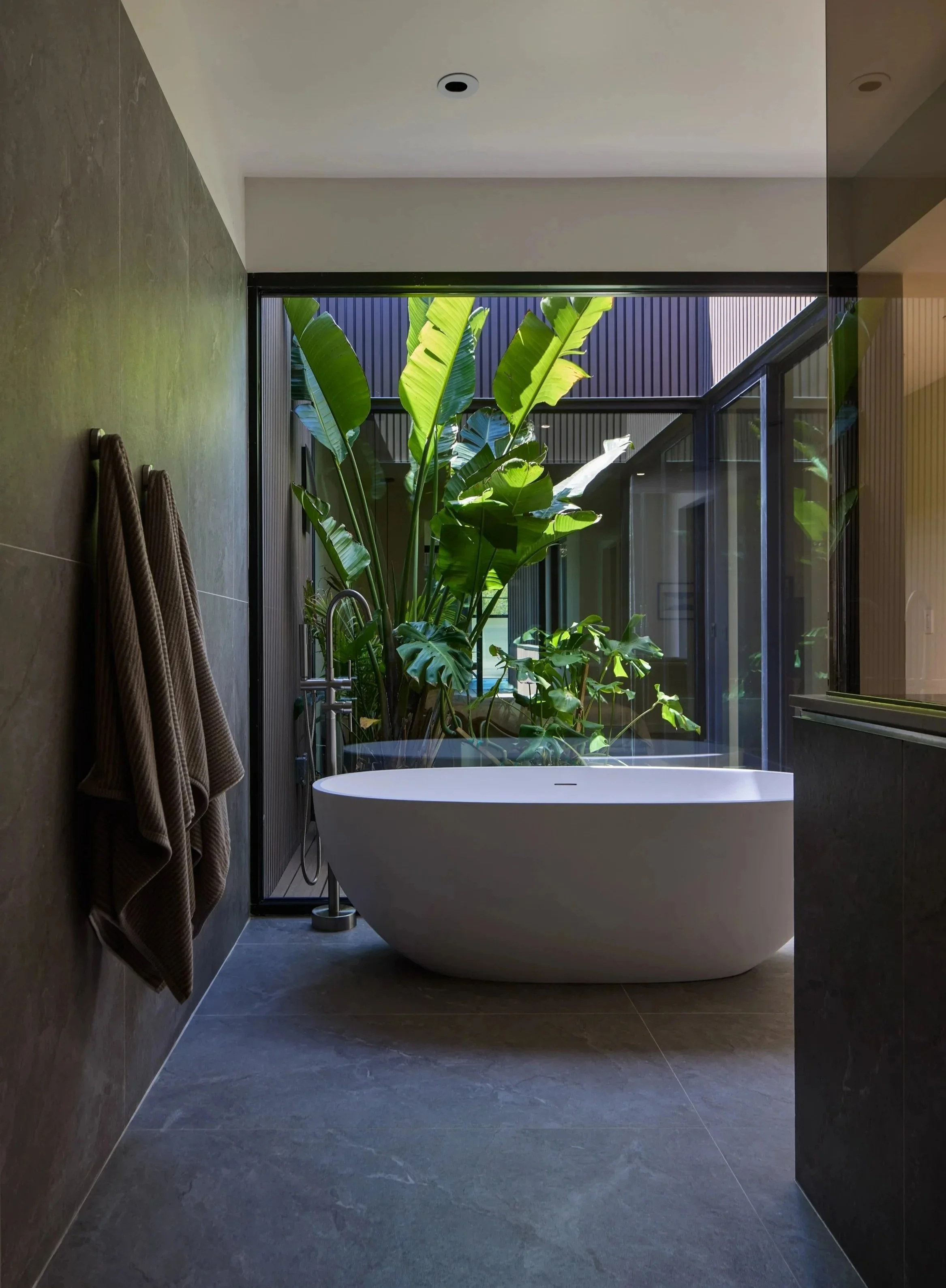 Modern bathroom with gray tiled flooring and walls, a freestanding white bathtub, large windows with greenery outside, and beige towels hanging on the wall.