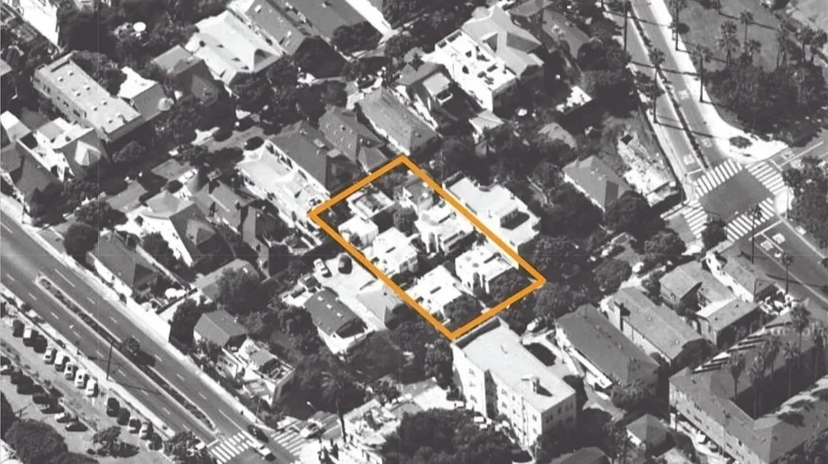 Aerial view of a residential neighborhood in black and white with an orange outline around a specific property. The neighborhood includes houses, trees, and streets with crosswalks. The outlined property shows a cluster of houses with driveways and p
