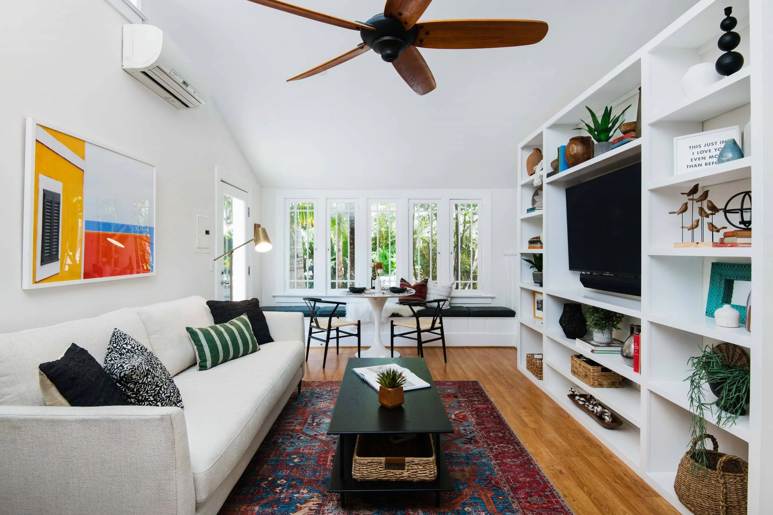 A cozy living room with white walls, a white sofa with black and green patterned pillows, a dark coffee table with a small potted plant, a multi-colored rug, a built-in white shelving unit with decorative items and books, a wall-mounted TV, a round d