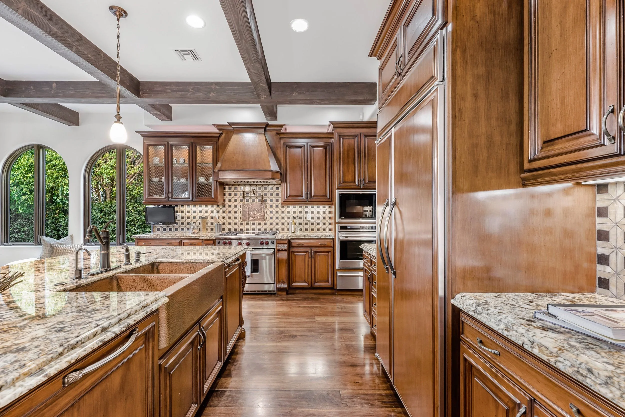 A spacious kitchen with wooden cabinets, a large granite island, and a tiled backsplash. Two arched windows reveal green foliage outside. Recessed lighting and a hanging pendant light illuminate the space.