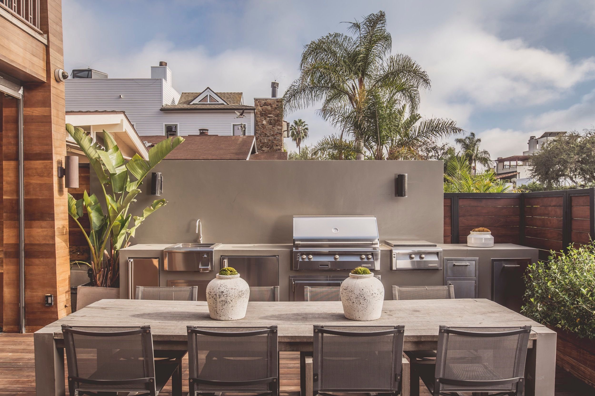 Outdoor patio with a dining table, chairs, potted plants, grill, and surrounding greenery including tall palm trees and plants.