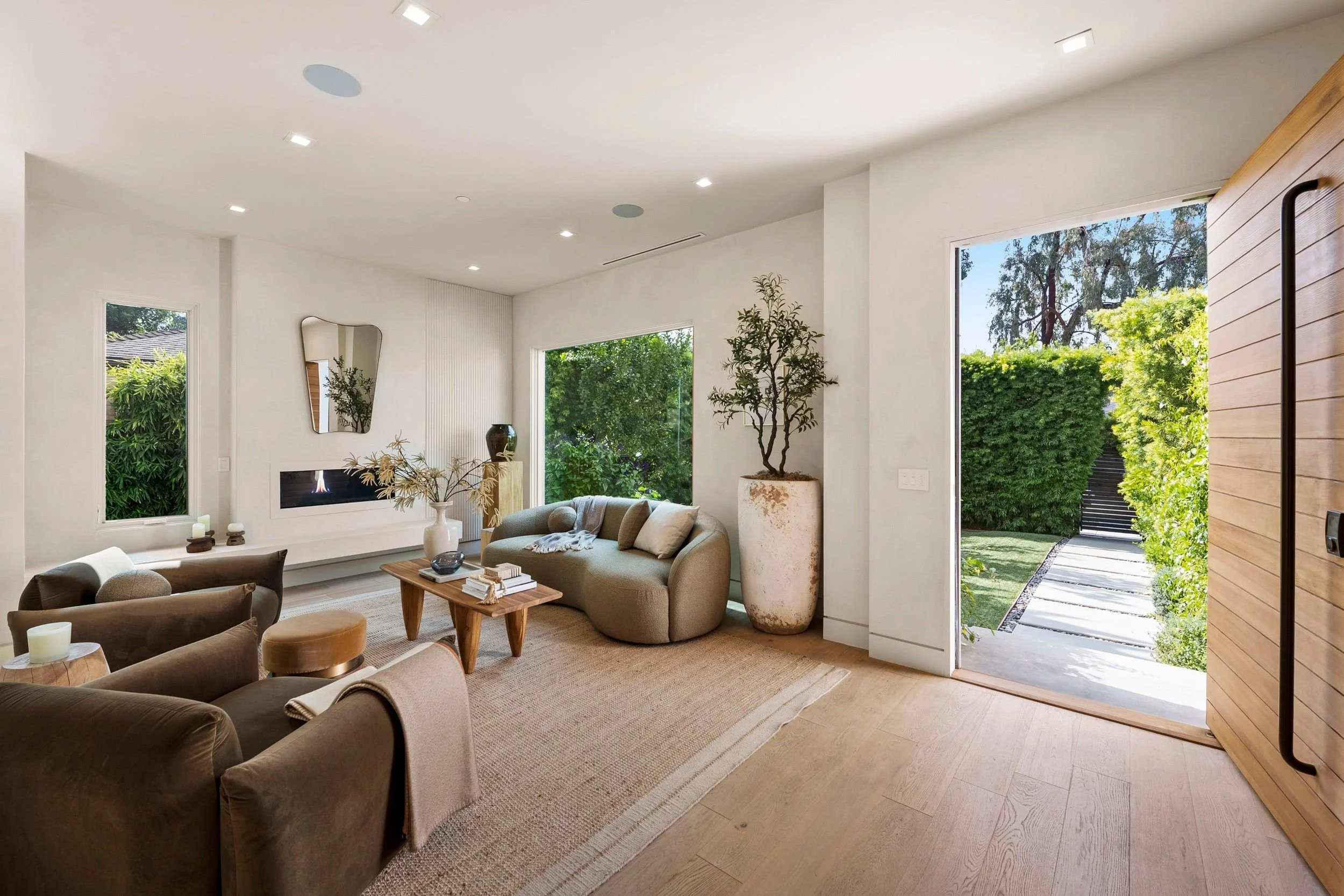 Living room with beige sofas, a wooden coffee table, large windows with greenery outside, and a door leading to a garden.