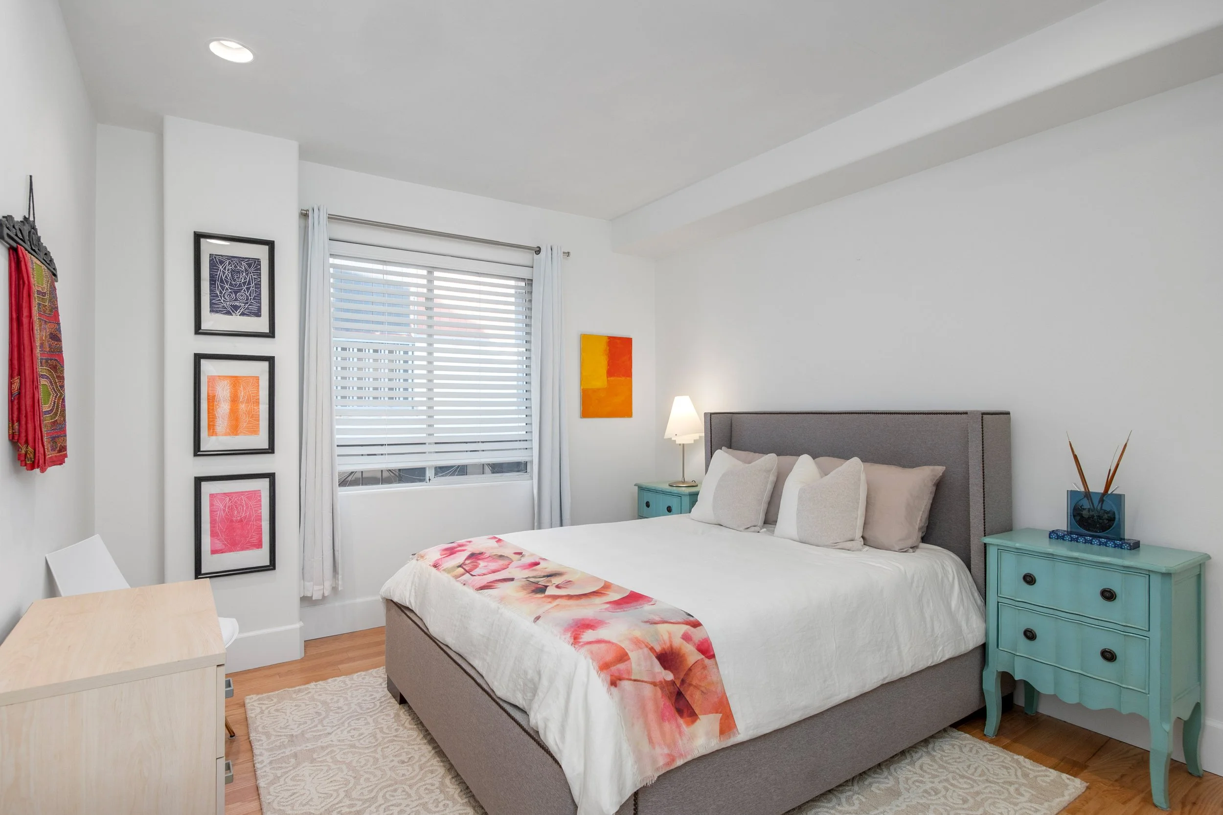 A bedroom with a bed that has beige pillows and a white blanket. Next to the bed is a light blue nightstand with dark drawer handles, holding a blue vase with greenery. The room has white walls, a window with white blinds and curtains, and colorful a