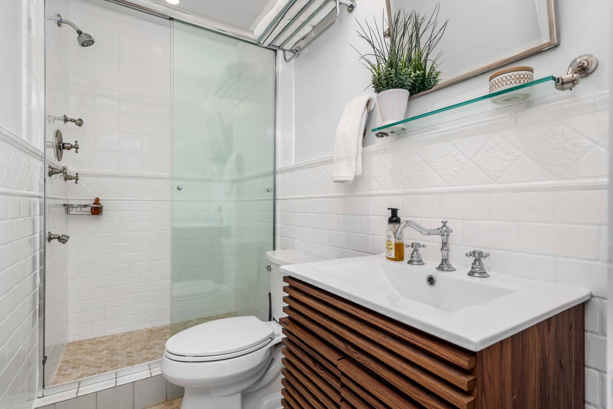 A bathroom with a walk-in shower with a glass door, white tiled walls, and a corner shower head. The bathroom has a white toilet, a vanity with a white sink, a dark wood slatted cabinet, a mirror, a potted plant, a towel, and shelves with a decorativ
