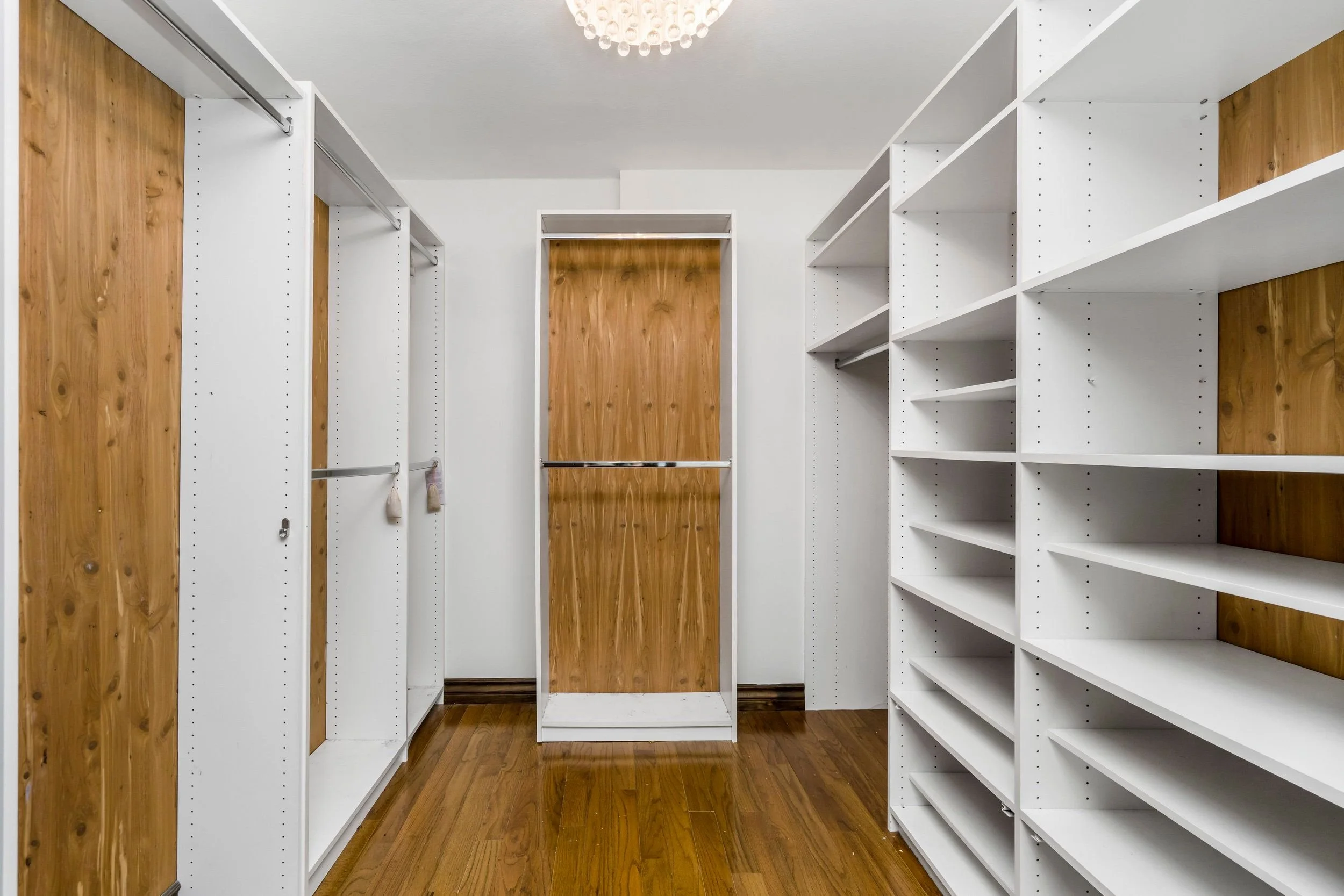 Empty walk-in closet with white shelves and wooden doors, hardwood floor, and a ceiling light fixture.