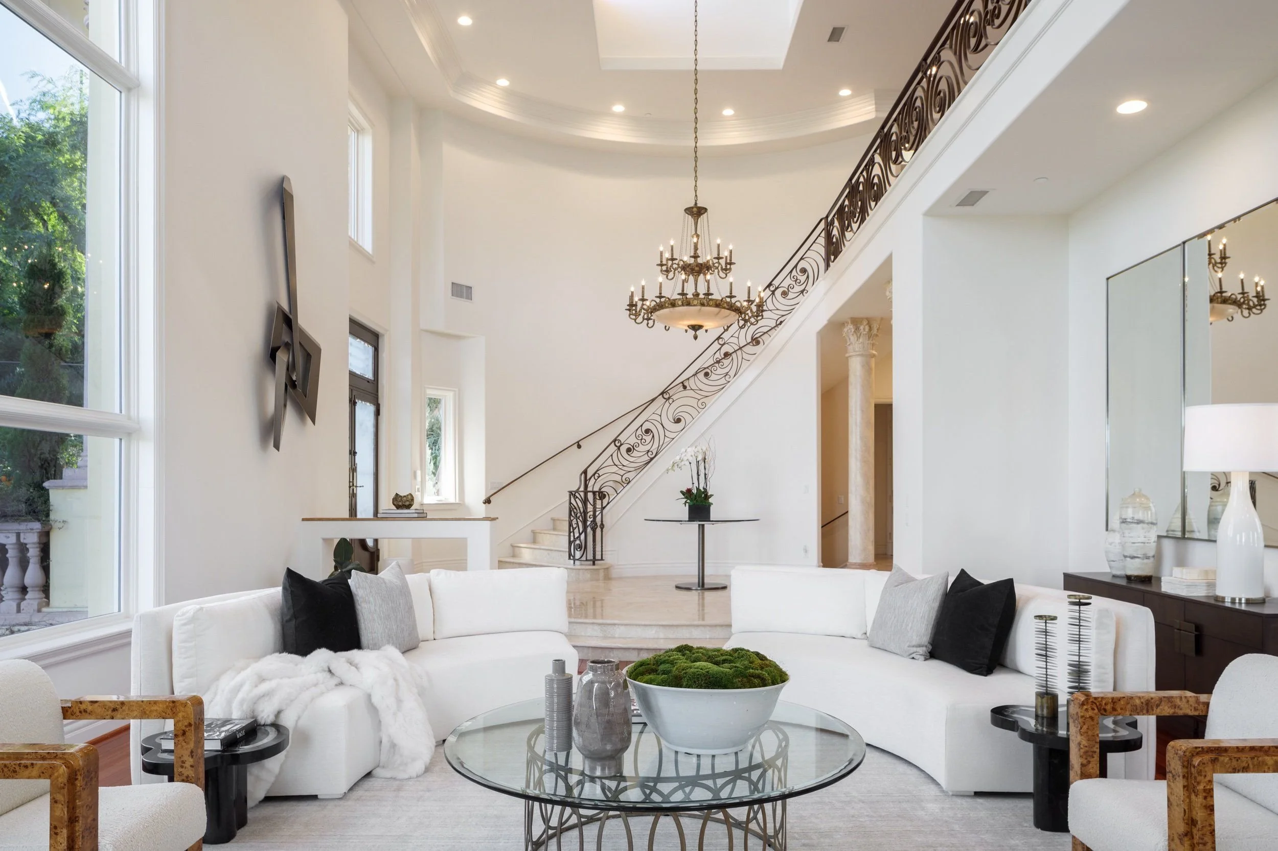 Modern living room with white sofa, glass coffee table, large windows, grand chandelier, and staircase with ornate railing.