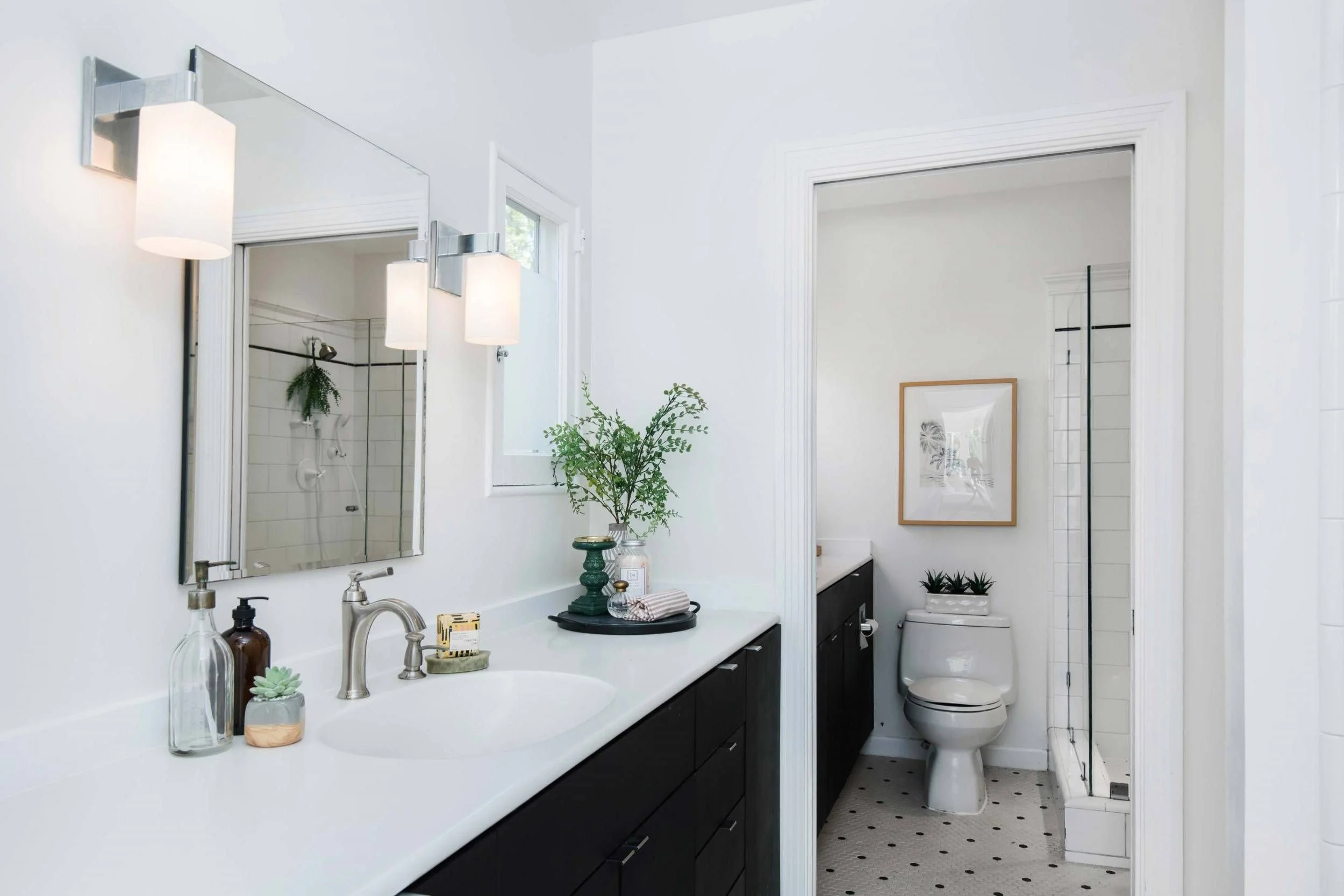 A modern bathroom with white walls, a large mirror, a white countertop with a sink, dark cabinets, and a toilet. Decor includes potted plants, candles, and framed art. There is a shower visible in the background with a glass door.