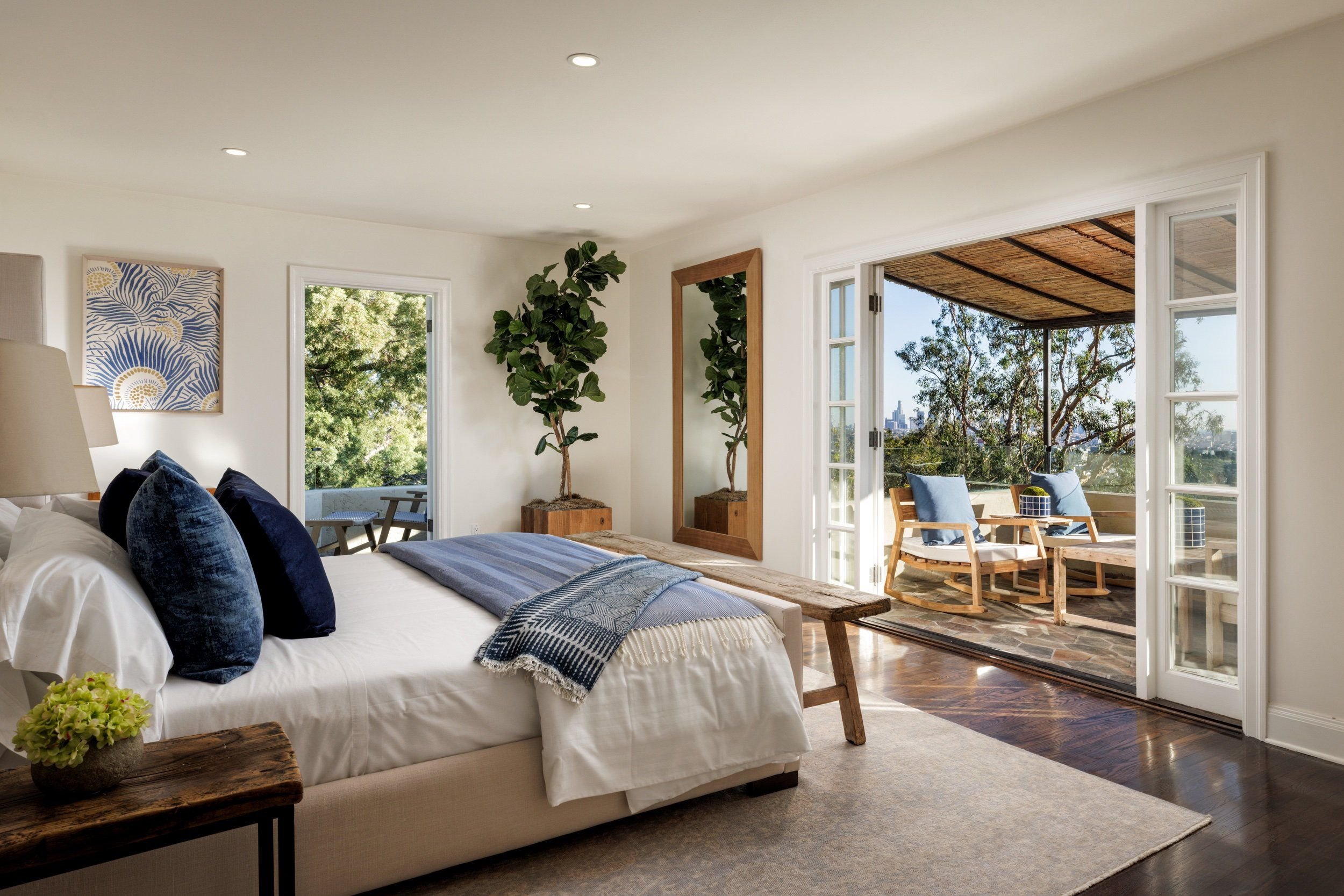 Bright bedroom with large windows and a balcony. The bed has white linens and navy pillows, with a wooden bench at the foot. There is a painting on the wall, a potted plant, and a small side table with a green floral arrangement. The balcony has wood