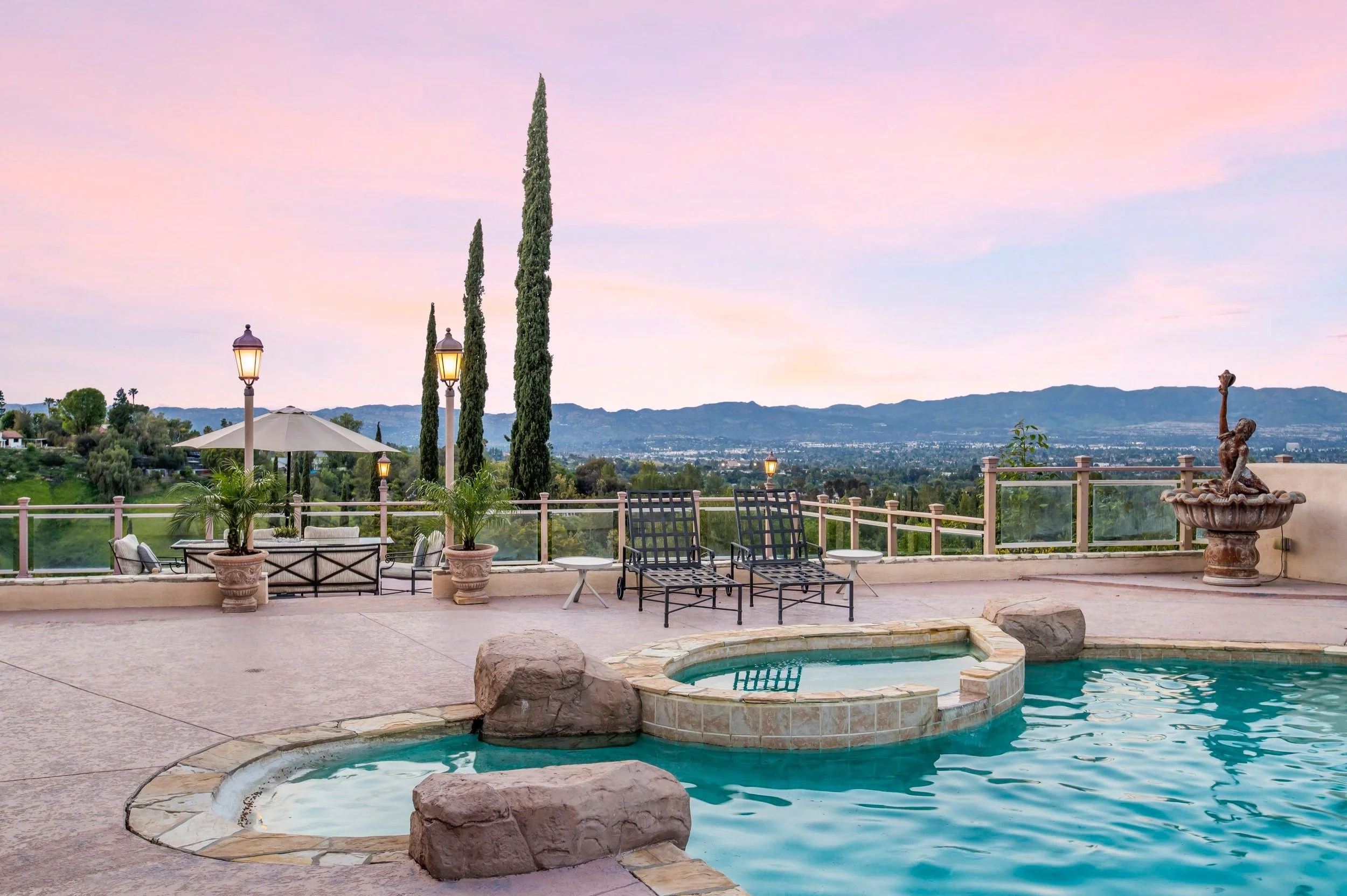 A backyard patio with a small swimming pool, lounge chairs, potted plants, and a wrought iron statue of a girl with a dolphin, overlooking a scenic view of rolling hills and a sky with pink and purple hues at sunset.