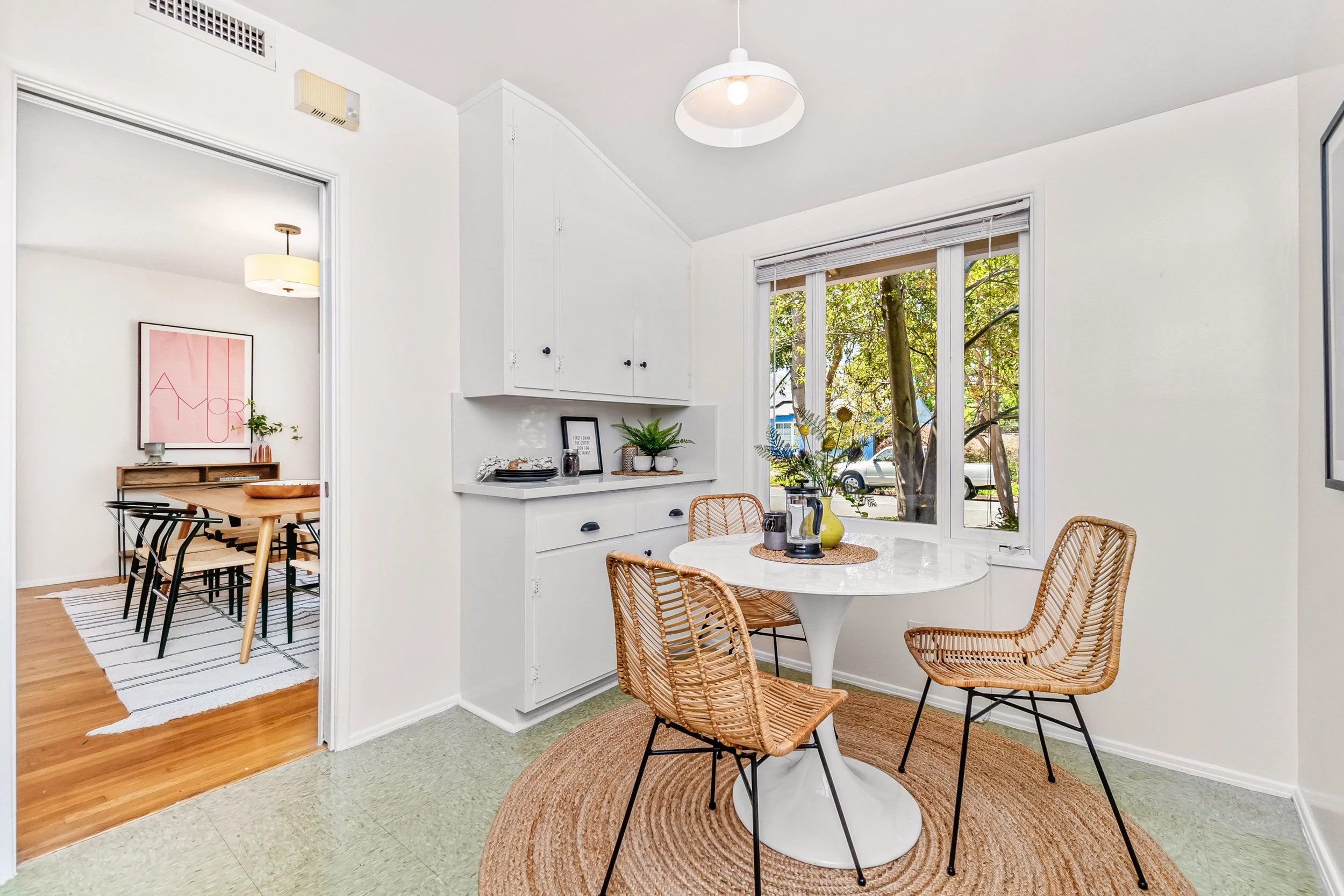 A dining area with a white round table surrounded by four rattan chairs, a small rug underneath, and a large window showing trees outside. The room has white walls and ceiling, with a modern hanging light fixture.