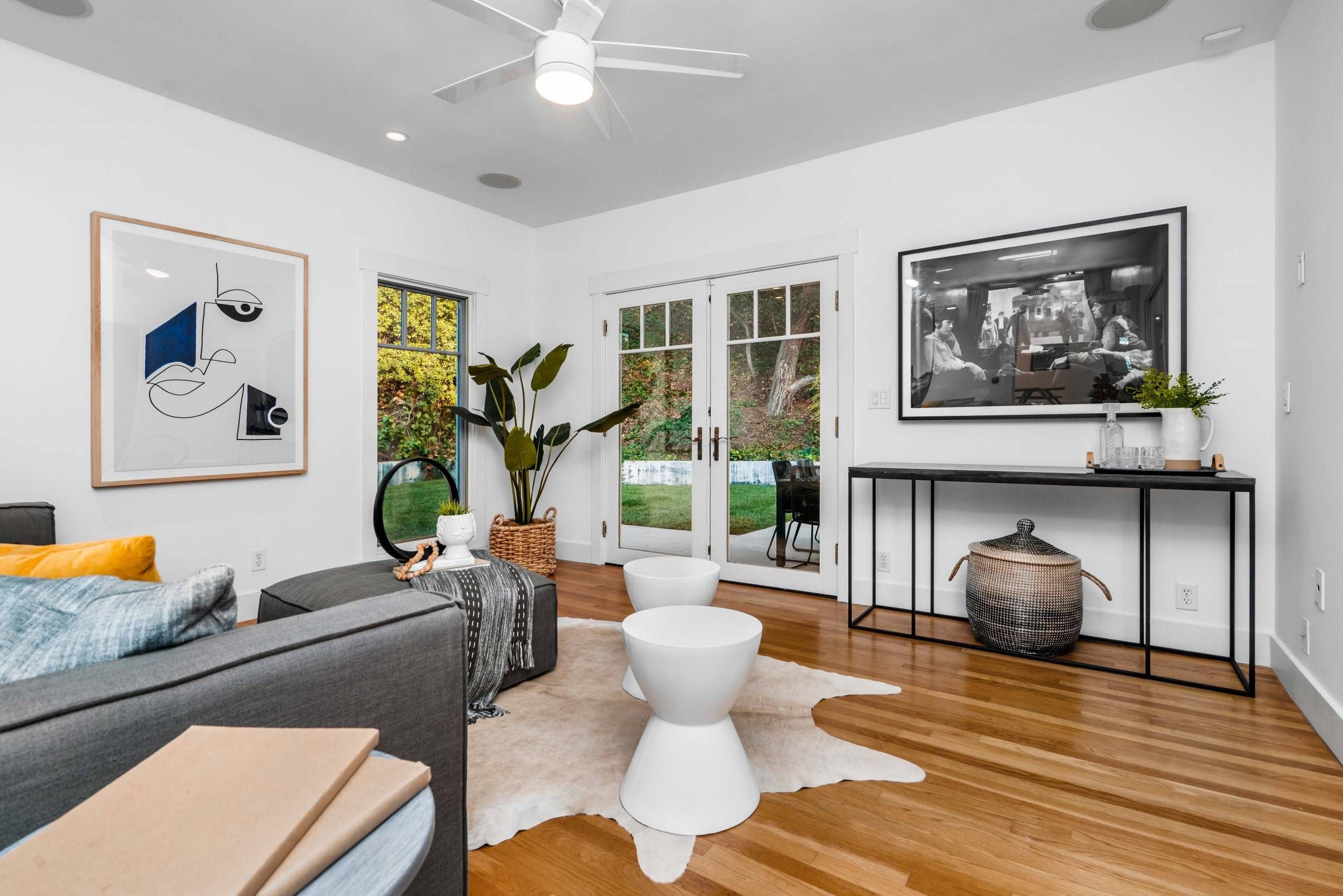 A living room with a gray sofa, white two-tiered tables, a wicker basket, wall art, a large plant, and a black console table with a potted plant, overlooking a backyard through glass doors.