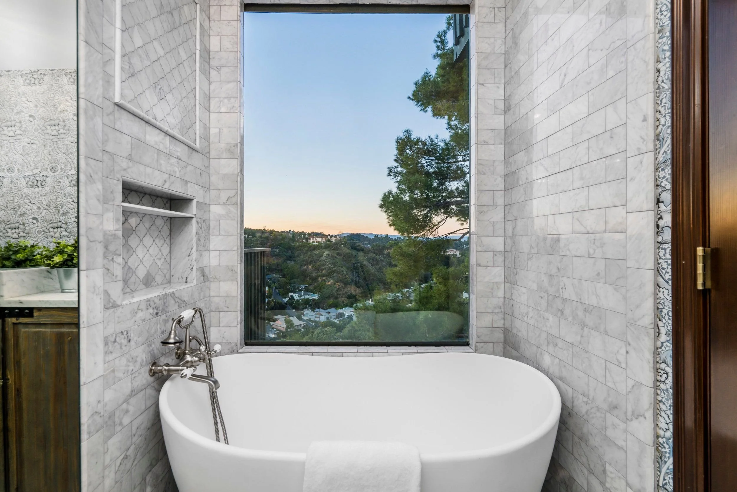 Bathroom with a bathtub by a large window showing trees and a distant view of houses and hills at sunset.
