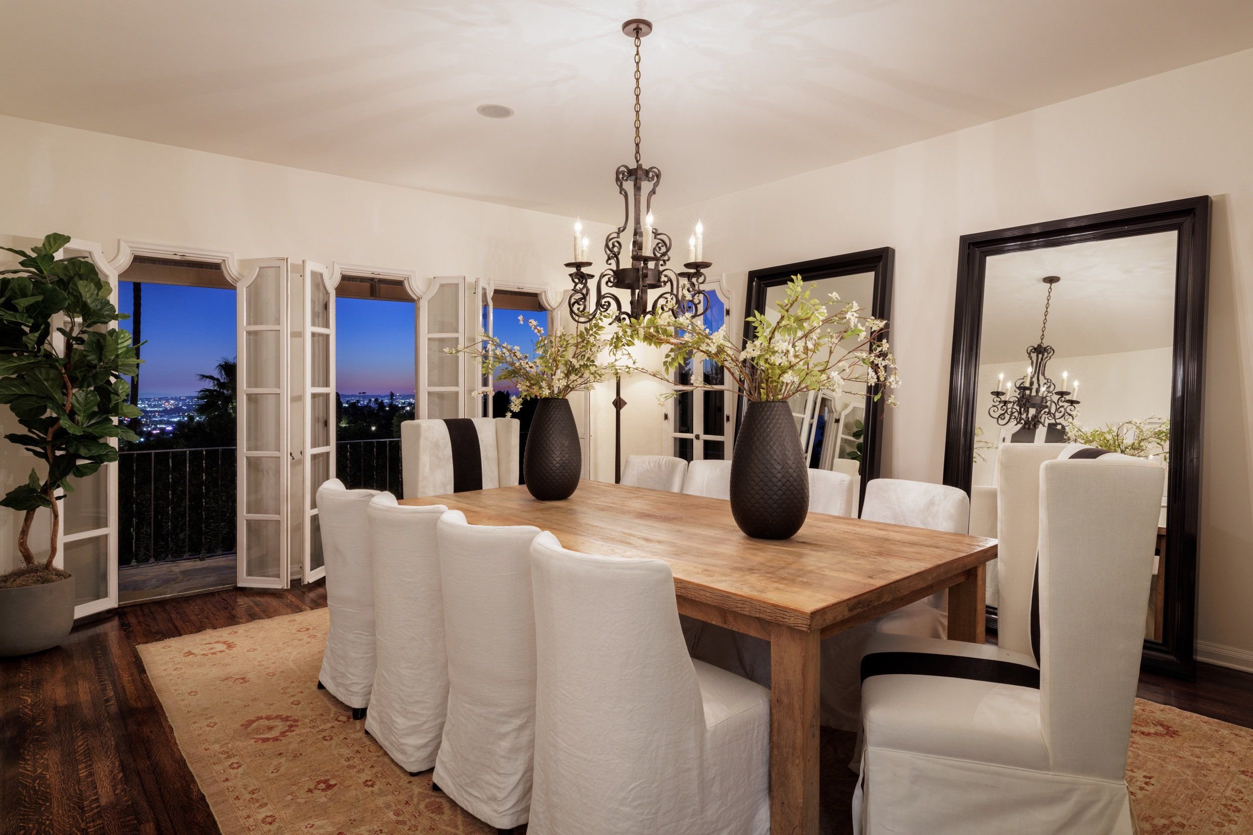 Elegant dining room with large wooden table, white upholstered chairs, two black vases with white floral arrangements, large black-framed mirror, chandelier, and open windows revealing a city view at dusk.