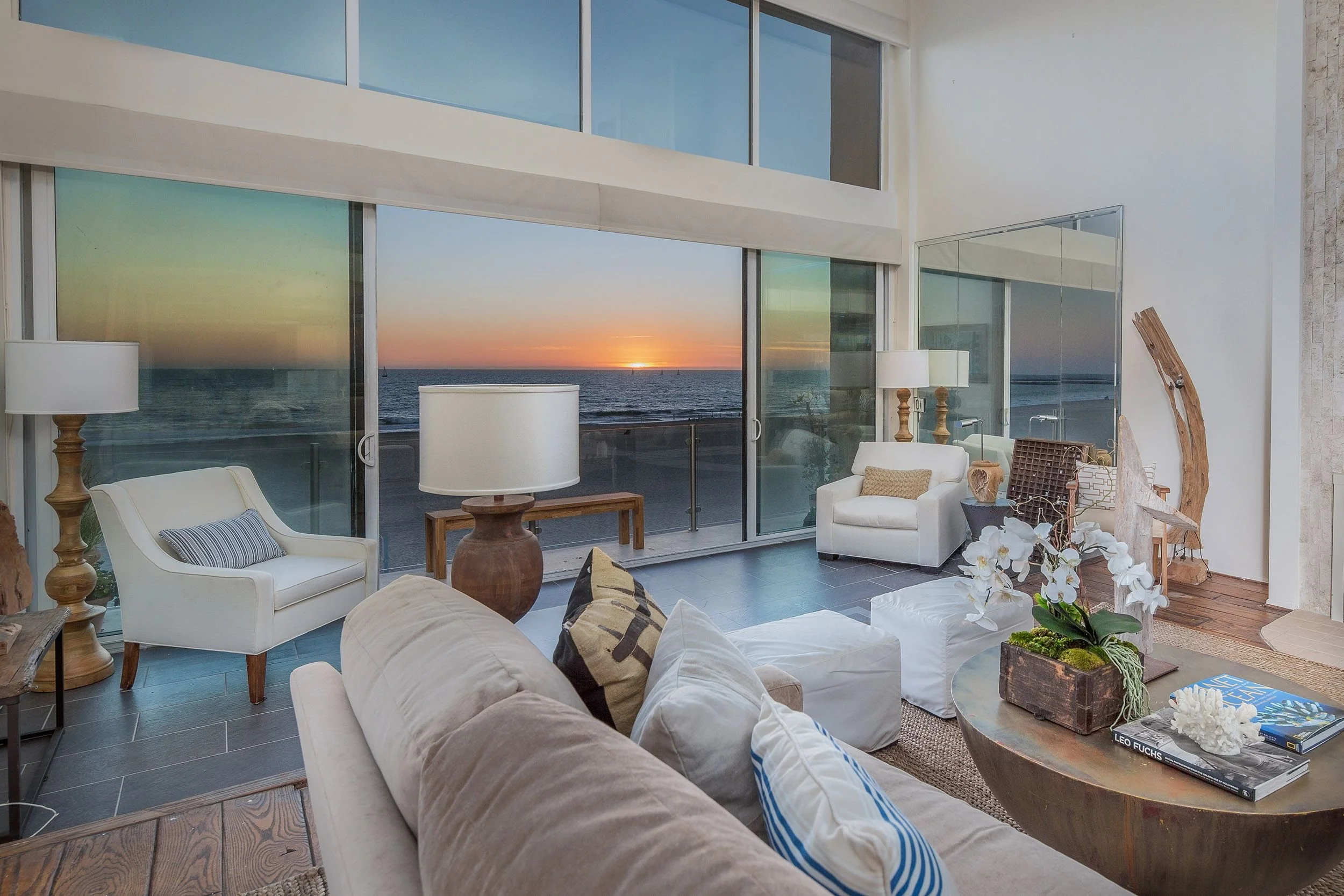 Living room with large windows showing ocean sunset view, white sofas, wooden coffee table with books and decorations, lamps, and indoor plants.