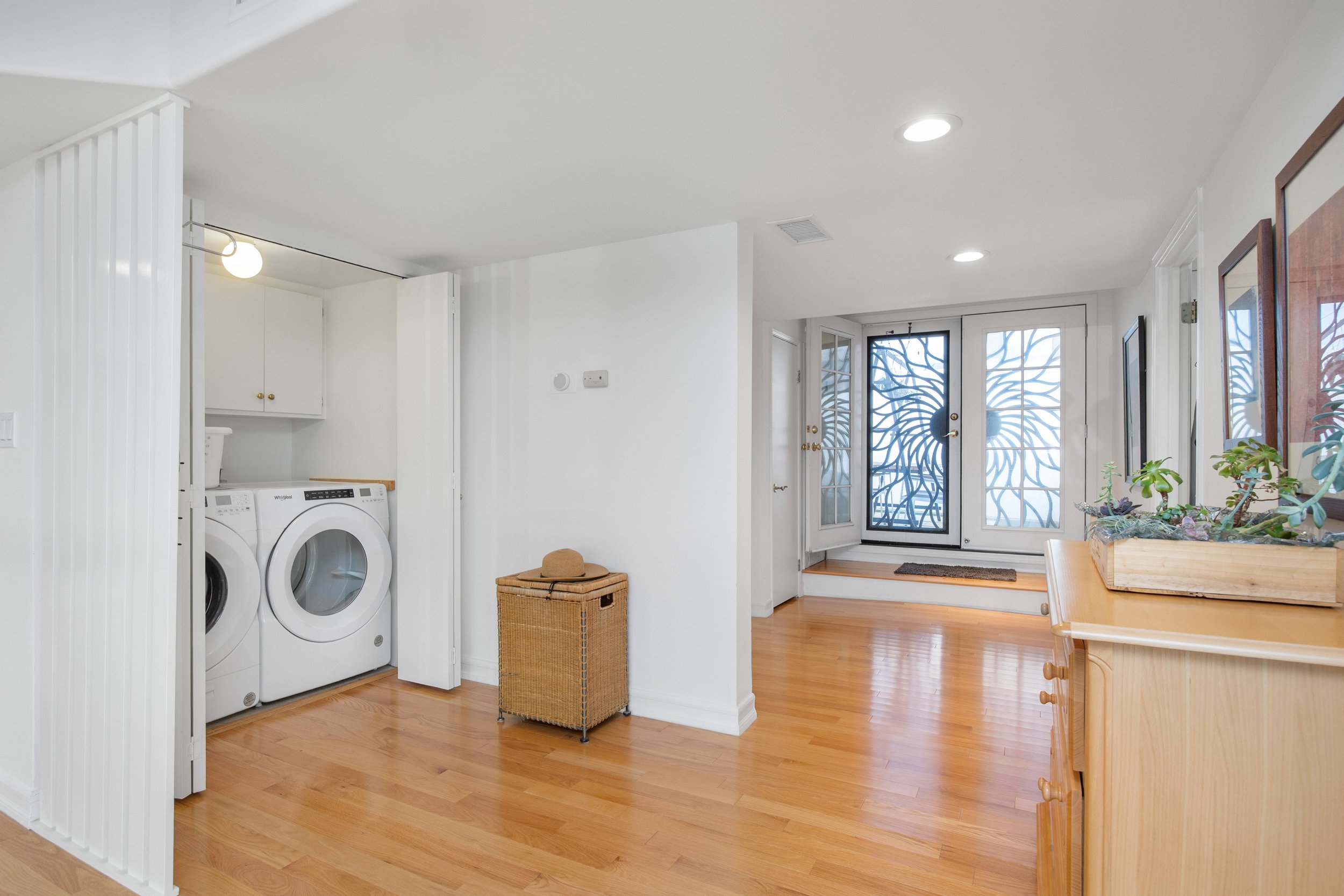Interior view of a laundry area with washer and dryer, a laundry basket, and a nearby door with decorative metalwork, leading outside. The space has wooden floors, white walls, and ceiling, with some framed artwork on the wall.