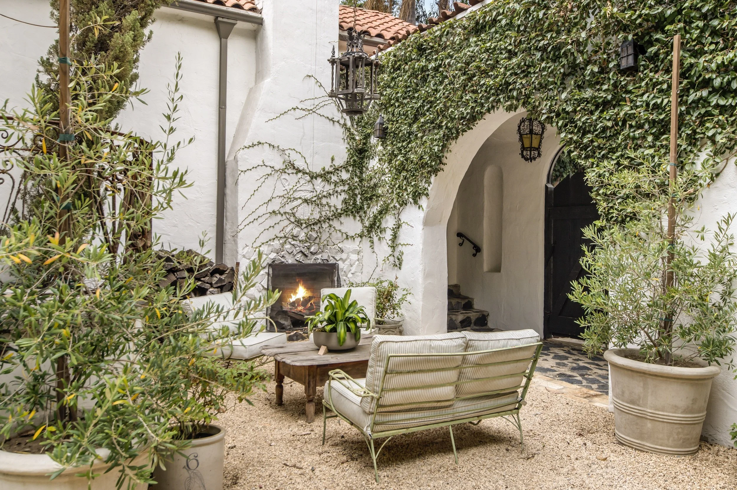 A cozy outdoor patio area with a fire pit, surrounded by comfortable seating and lush greenery, including potted plants and climbing vines on white walls.