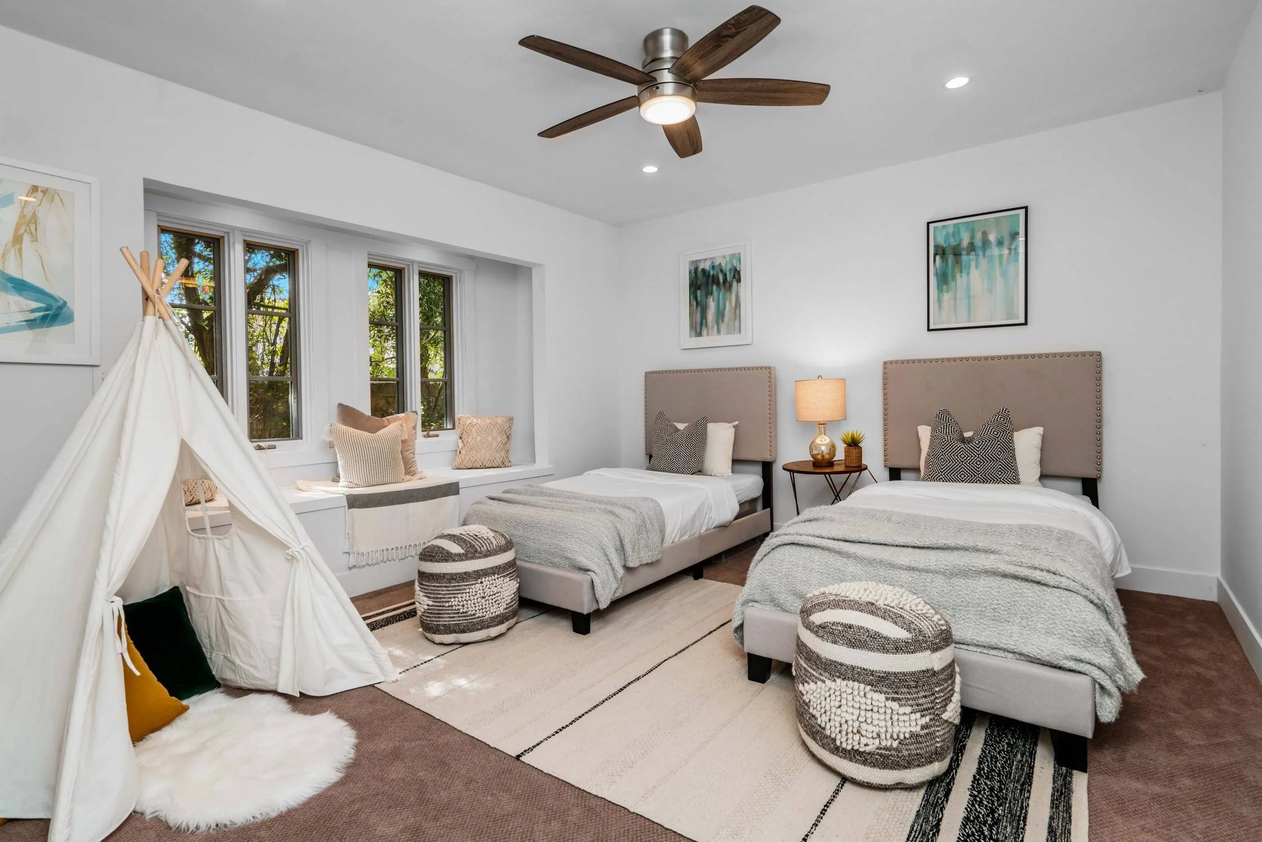 A bedroom with two beds featuring beige headboards, various patterned pillows, a nightstand with a lamp, framed abstract art on white walls, a ceiling fan with wooden blades, a row of three windows with a built-in window seat decorated with pillows, 
