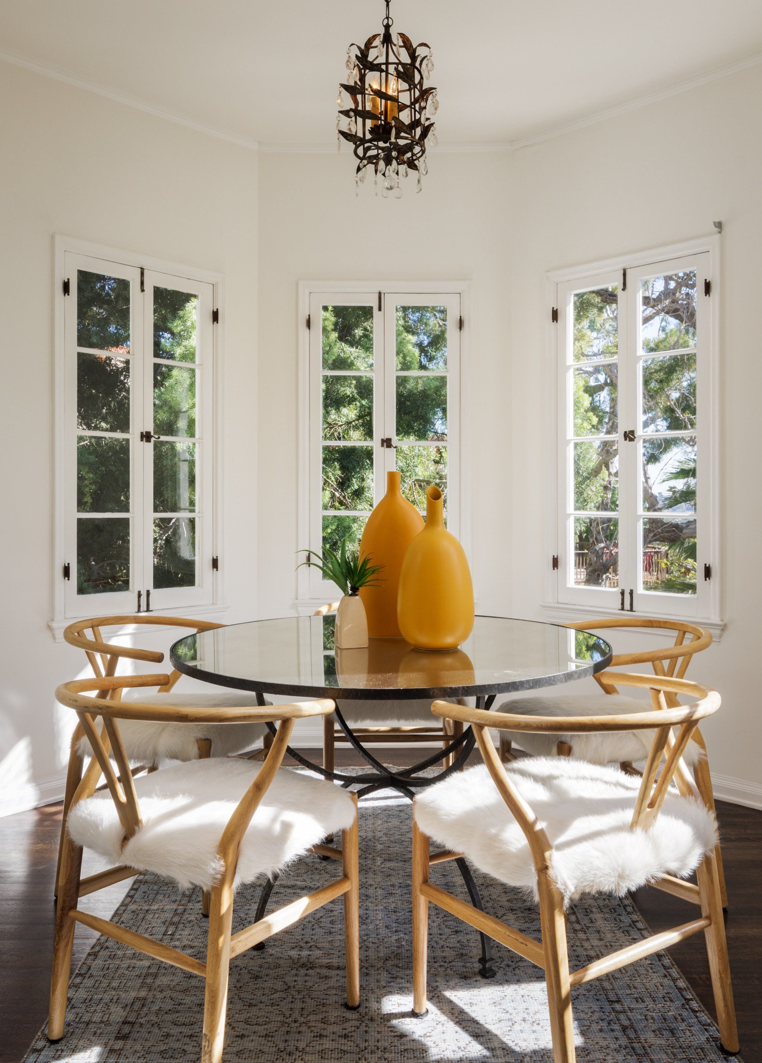 Sunlit dining room with white walls and large windows, round glass table with yellow vases, surrounded by six wooden chairs with white fur cushions, a chandelier hanging from the ceiling, and a rug under the table.