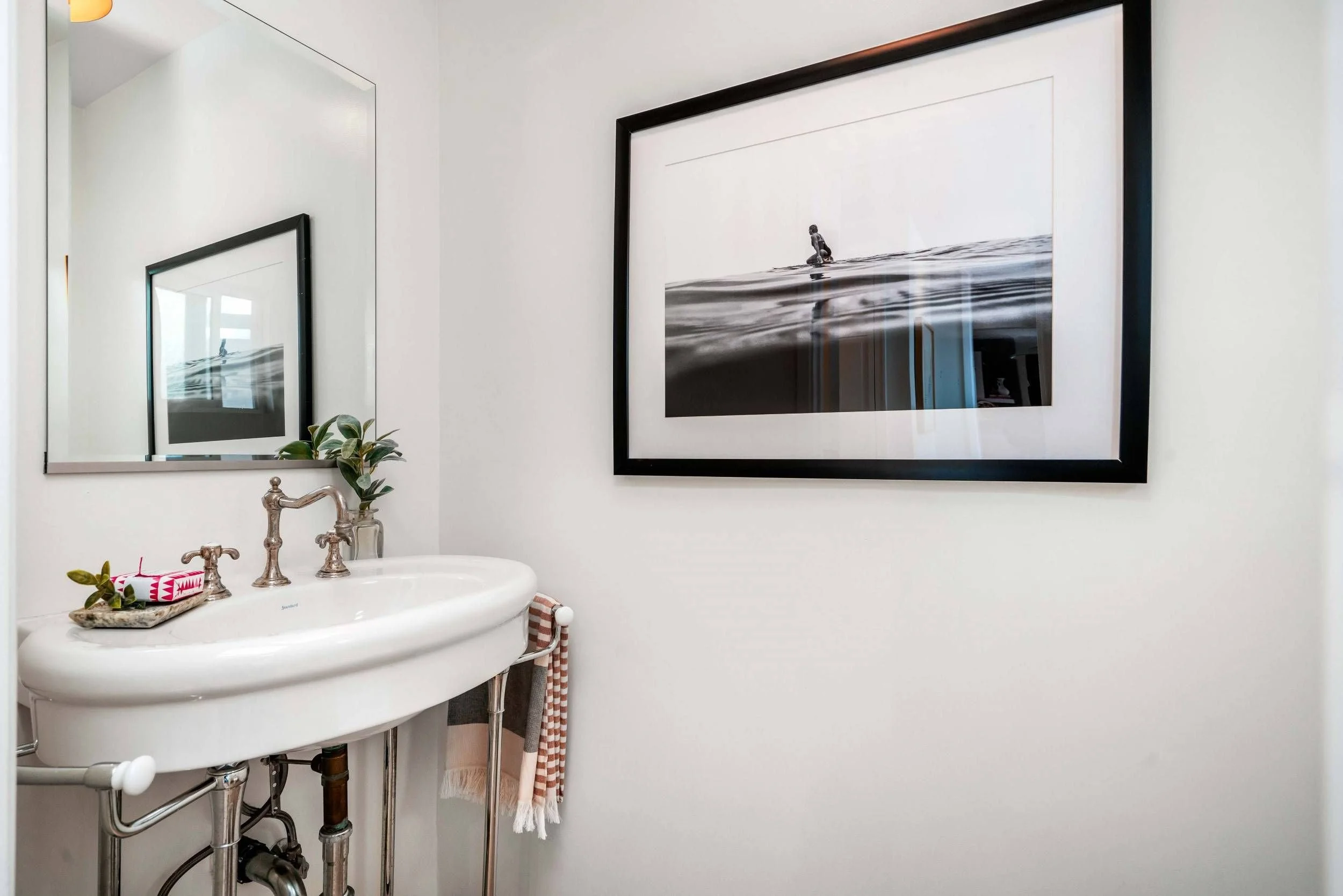 Bathroom with a vintage sink, a mirror, a framed black-and-white photograph of a surfer, a plant, and a striped towel hanging from a towel rack.