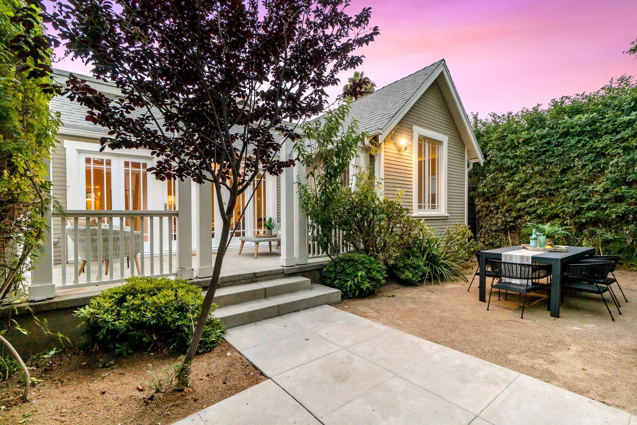 A backyard with a small patio, outdoor dining table with black chairs, lush green bushes, a tree, and a house with light-colored siding. The sky has a pinkish hue at dusk.