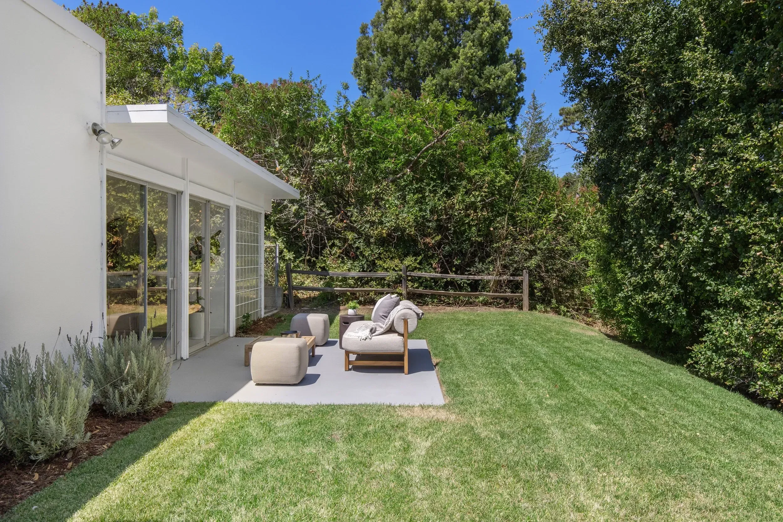 A backyard patio with outdoor seating, including cushioned chairs and a small table, adjacent to a white house with large windows. The patio extends to a grassy lawn surrounded by tall green trees and bushes under a clear blue sky.